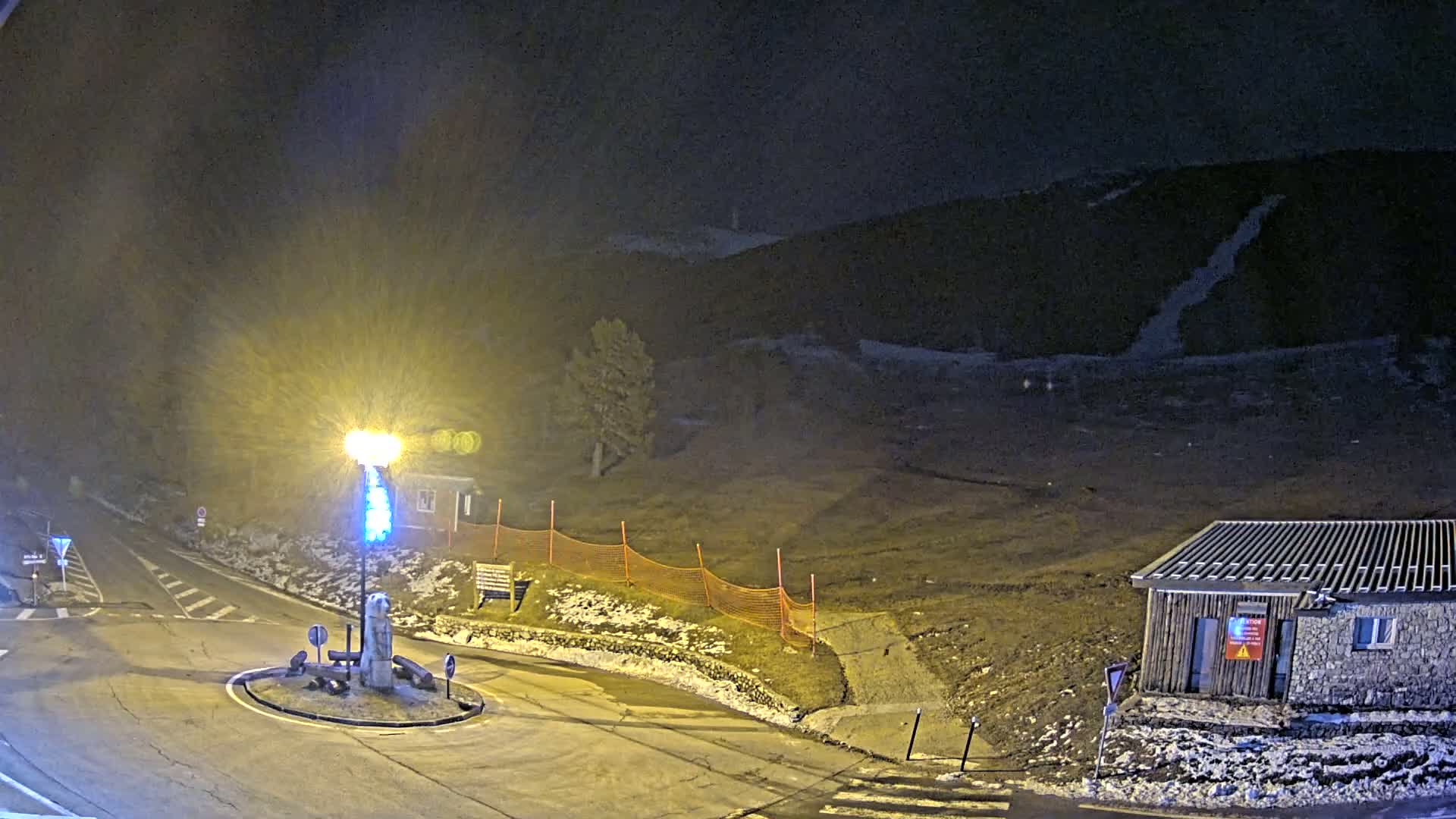 A nighttime view of a roundabout with a small building and a hill in the background, under a dark sky with some light snow on the ground.