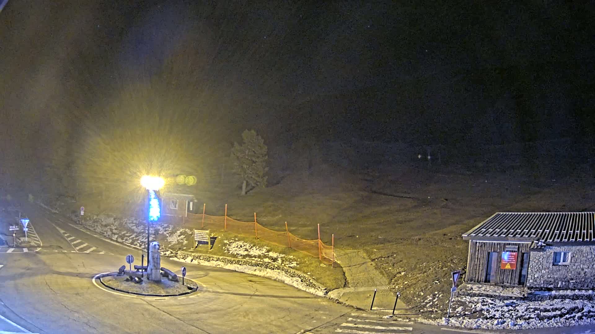 A brightly lit roundabout at night, partially covered in snow, features a small building and an orange construction fence.