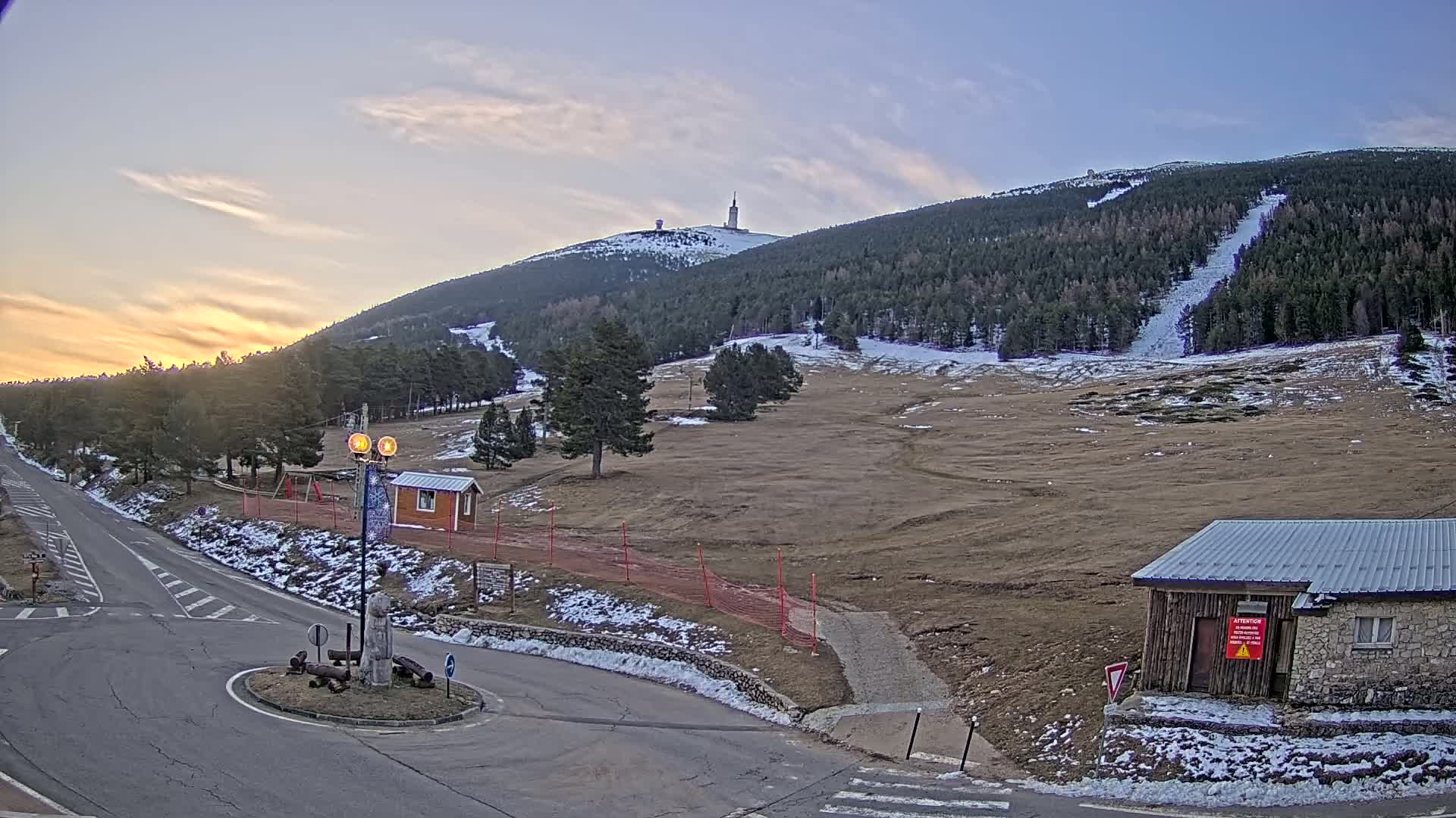 A mountaintop with a tower is visible in the distance under a partly cloudy sunset sky, with a road, small buildings, and patchy snow on the ground in the foreground.
