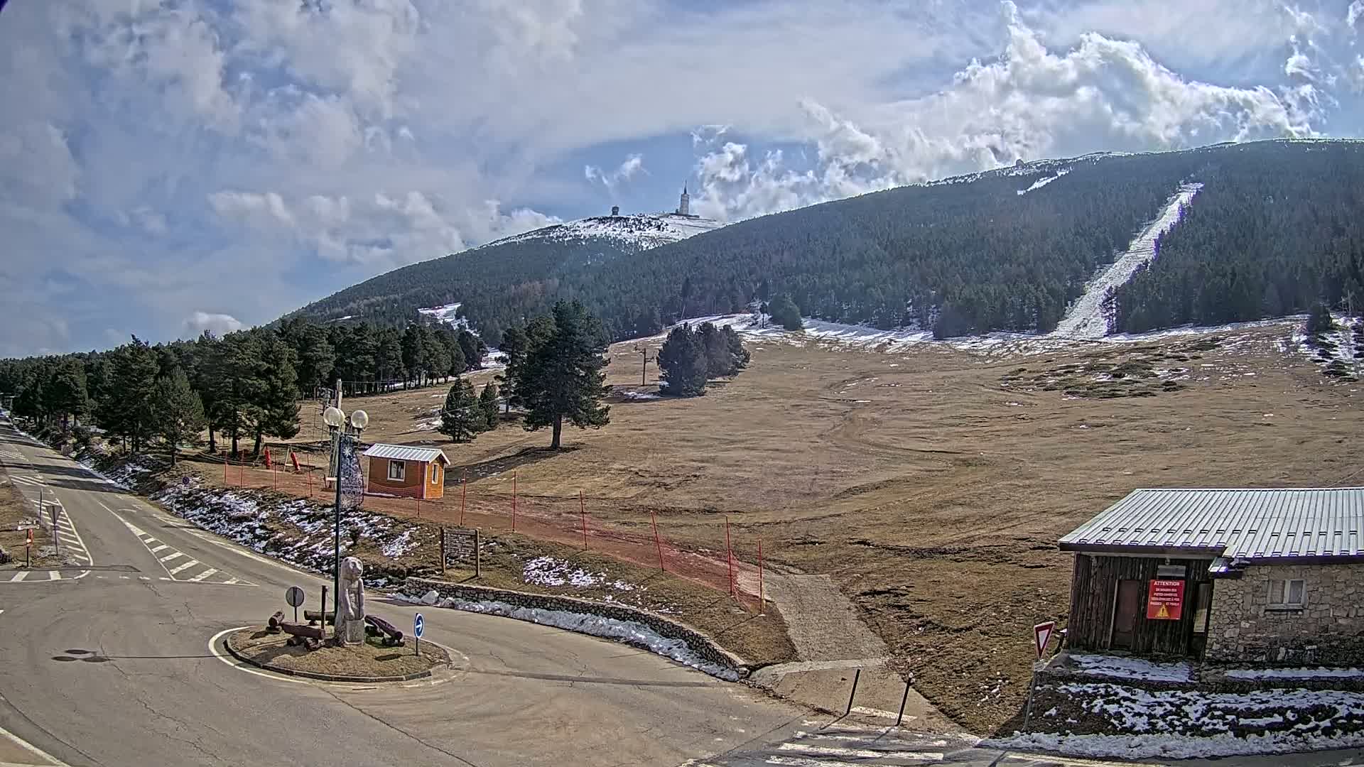 A mostly sunny day reveals a mountaintop with a communications tower, ski slopes, and surrounding coniferous forests, viewed from a paved road near a small building and roundabout.