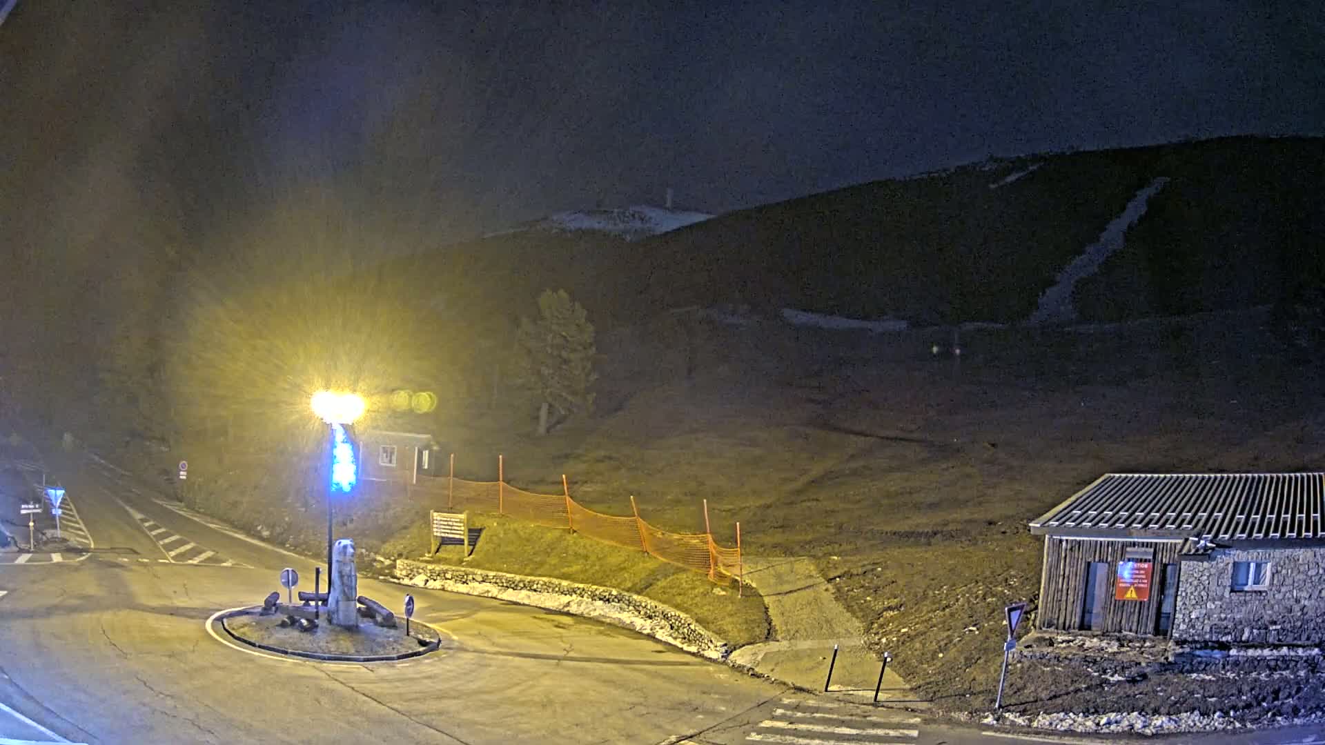A nighttime view of a roundabout with a small building and a hill in the background under dark, possibly snowy conditions.
