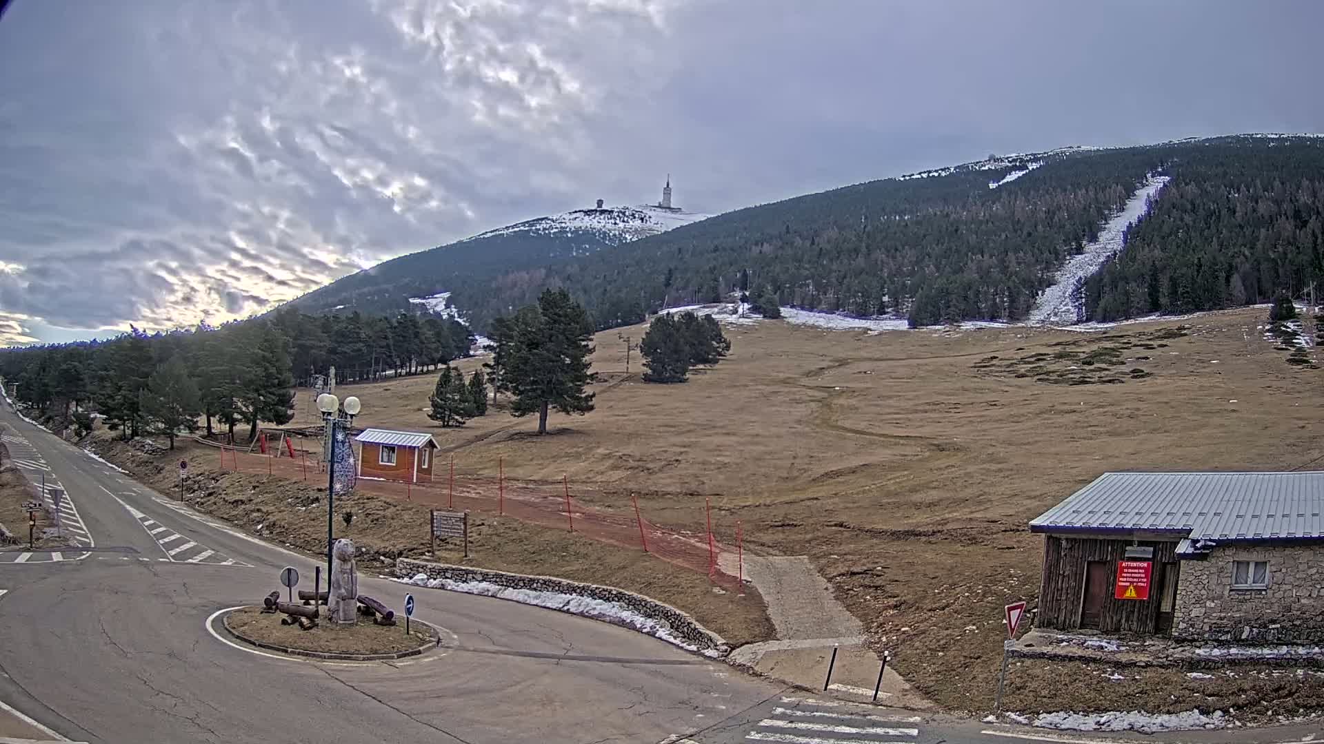 A mountaintop with a tall tower is visible in the distance under a partly cloudy sky, while a road leads to a roundabout near some small buildings in the foreground, with patches of snow on the ground.