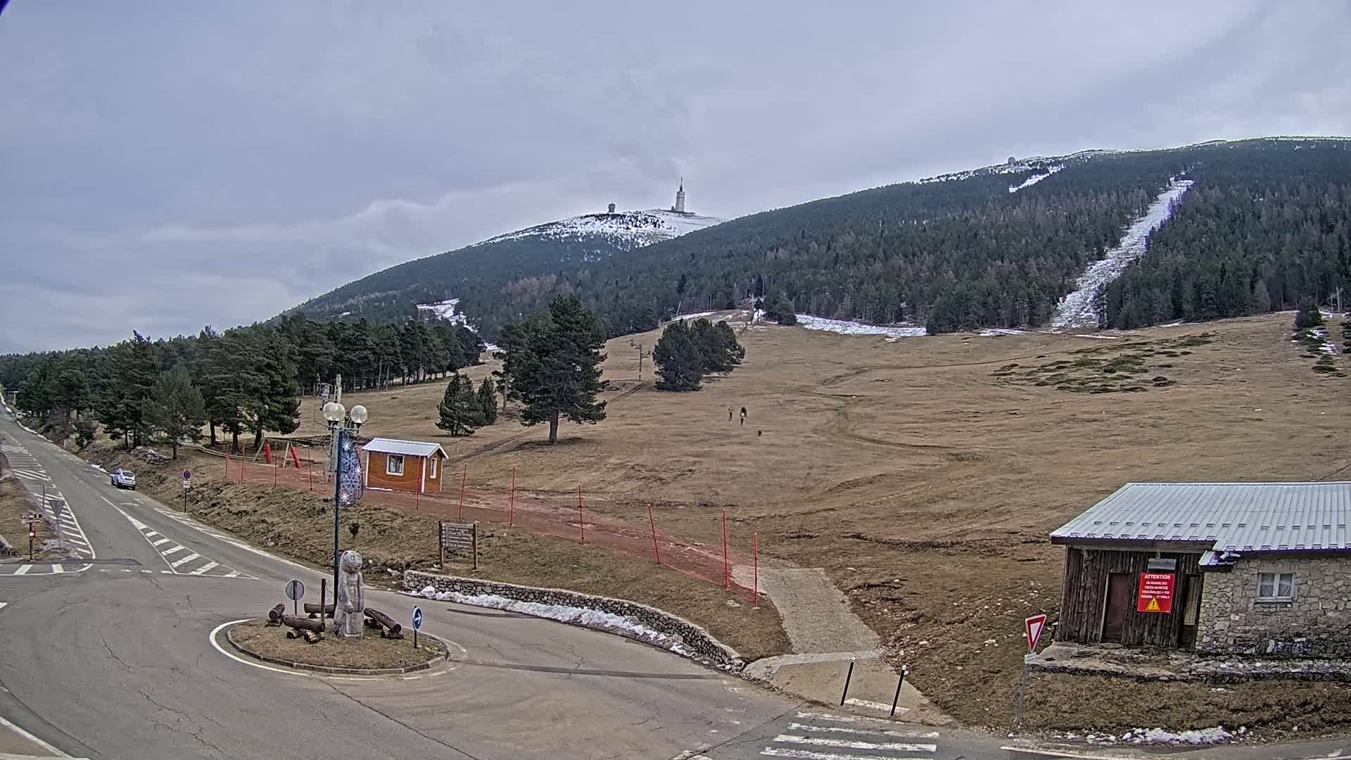 A mountaintop with a tall tower is visible in the distance under a cloudy sky, with a sparsely populated, dry, mostly brown hillside in the foreground featuring a road, small buildings, and a few people.