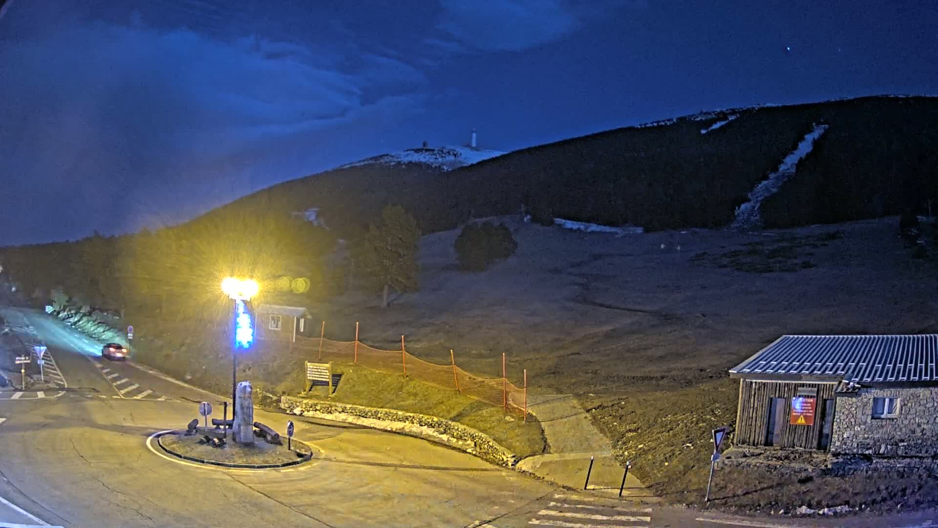 A nighttime view of a snow-dusted hill with a small building and a roundabout, illuminated by a streetlight under a dark blue, clear sky.