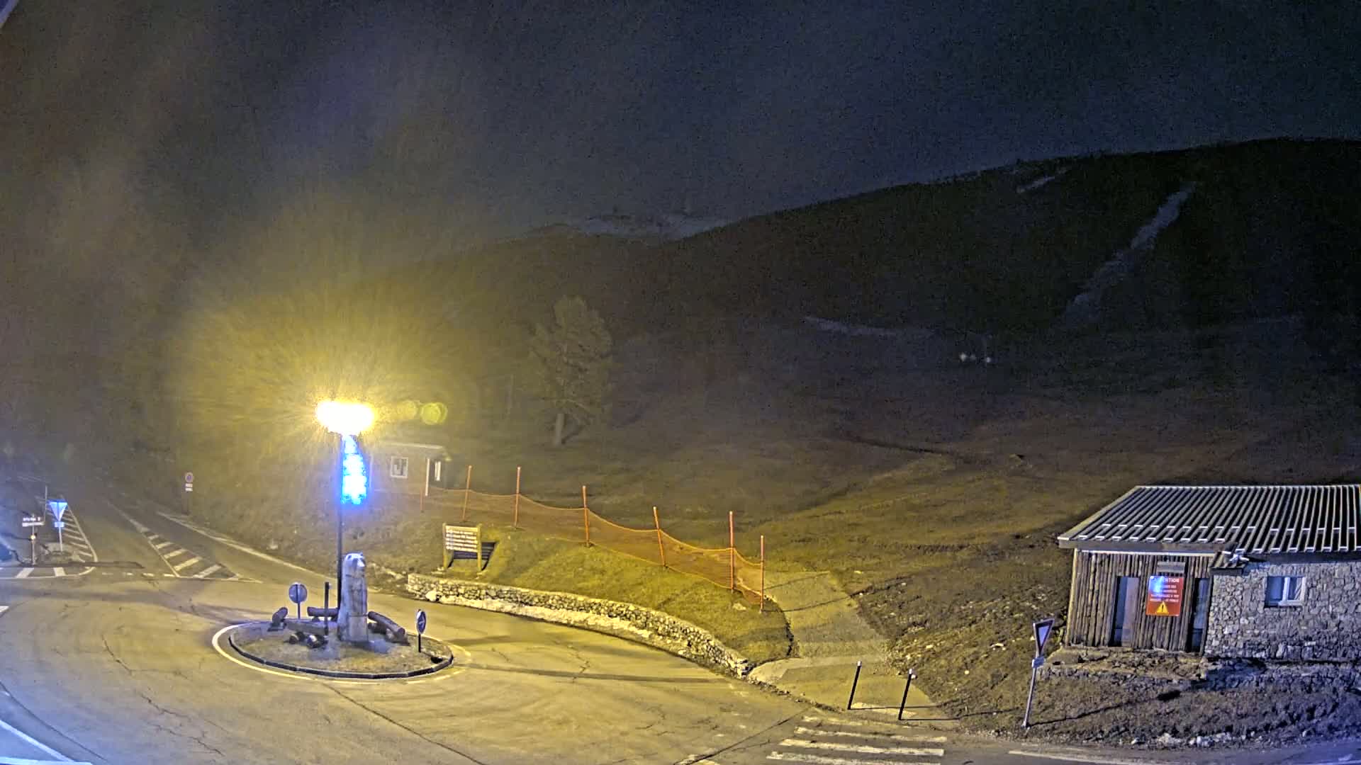 At night, a brightly lit roundabout sits in front of a small building and a hill under hazy, dark conditions.
