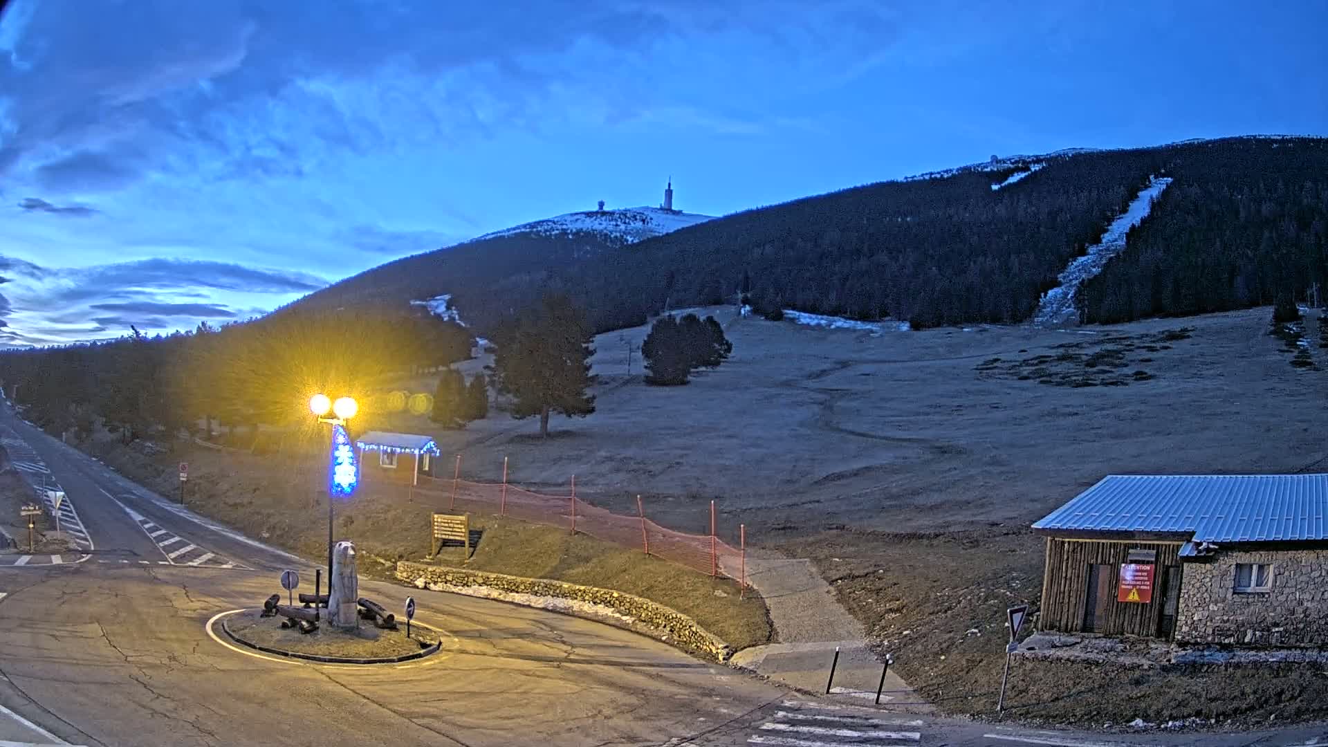 A mountaintop with a tall tower is visible in the distance under a twilight sky, with a streetlight illuminating a roundabout and small building in the foreground.