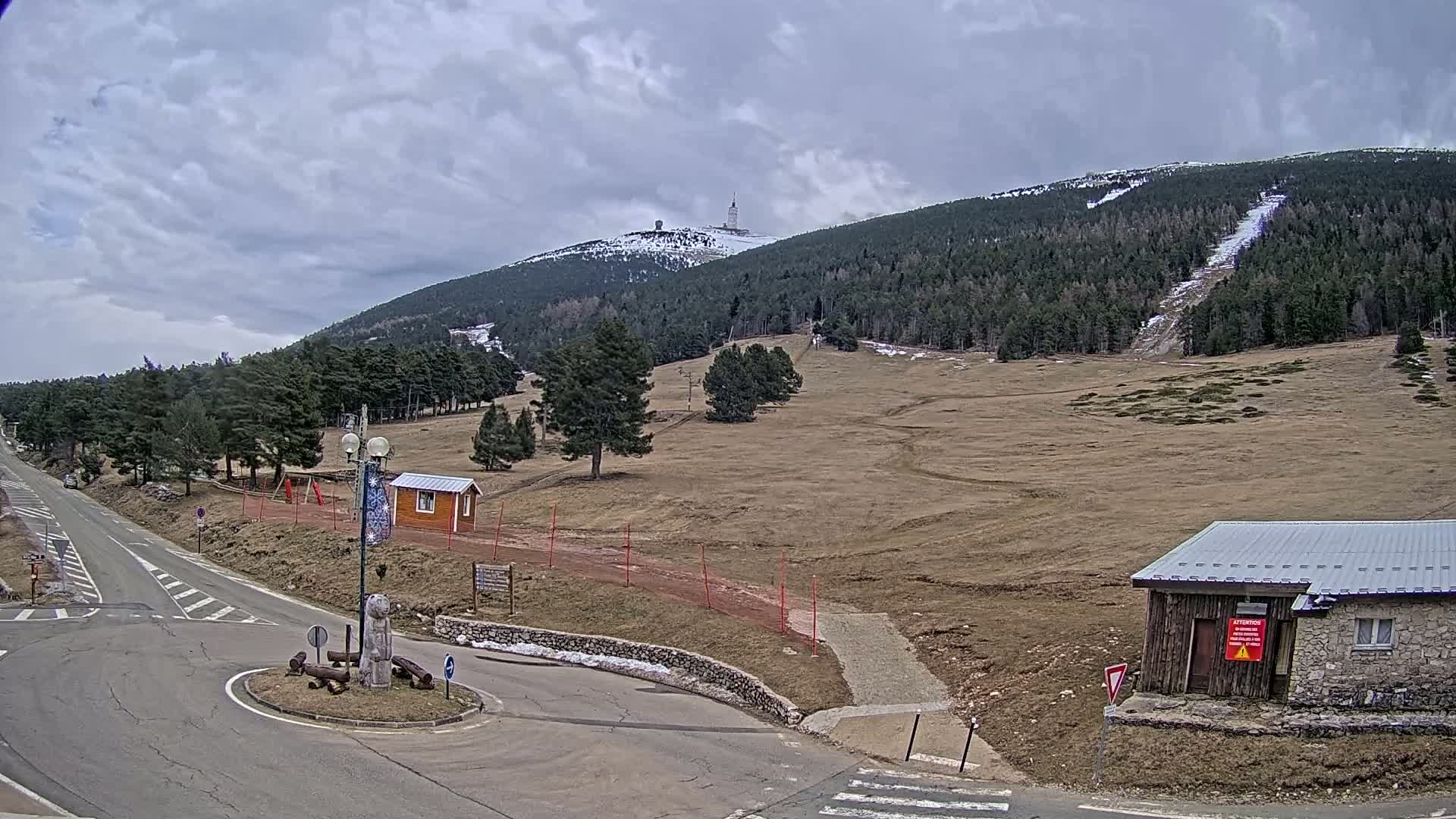A mountaintop with a tower is visible in the distance under a cloudy sky, overlooking a mostly barren landscape with some sparse trees, roads, and small buildings.