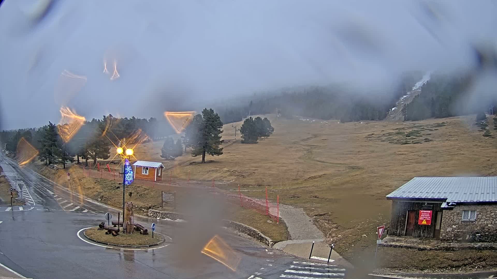 A wet, foggy scene shows a road intersection near a ski slope with sparse trees and small buildings, under a cloudy sky.