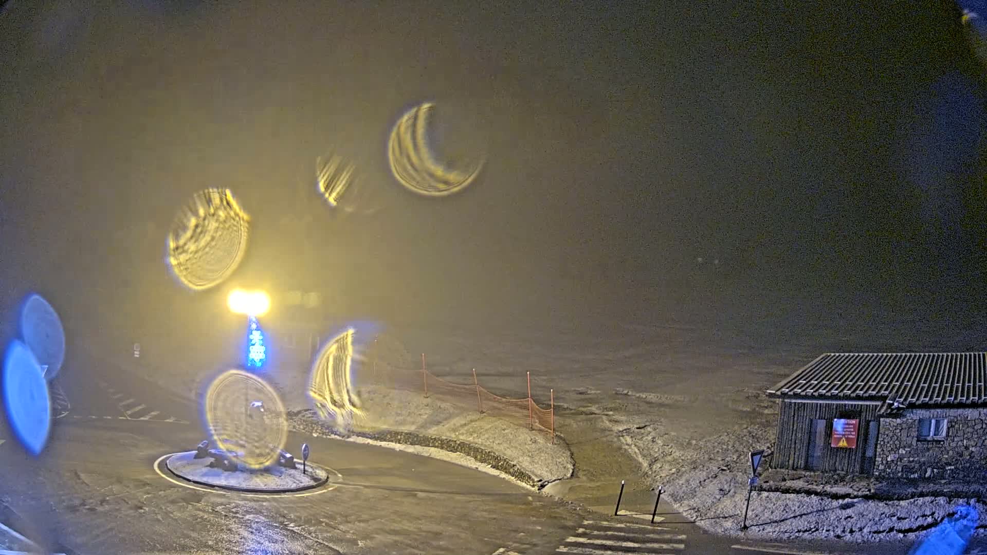A nighttime, snowy scene viewed through rain-covered glass shows a small building, a circular roadway with a light post, and a snow-covered area beyond.