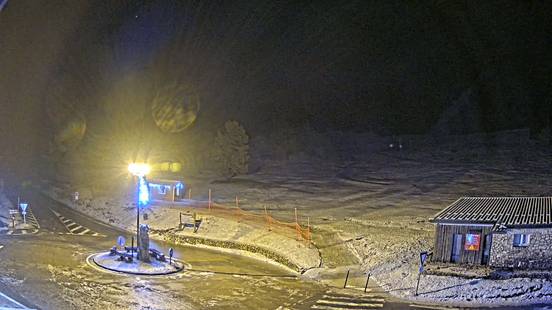 A snow-covered roundabout at night, illuminated by a streetlight, with a small building and orange safety netting in the background.