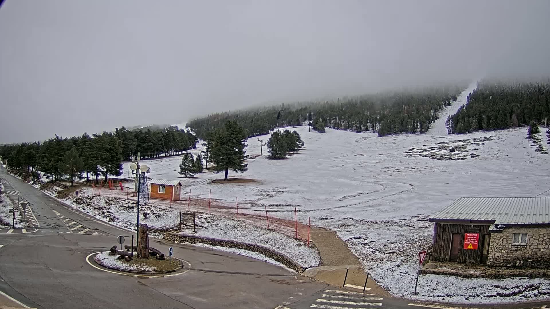 A snow-covered ski slope is seen from a roadside viewpoint, under an overcast sky.