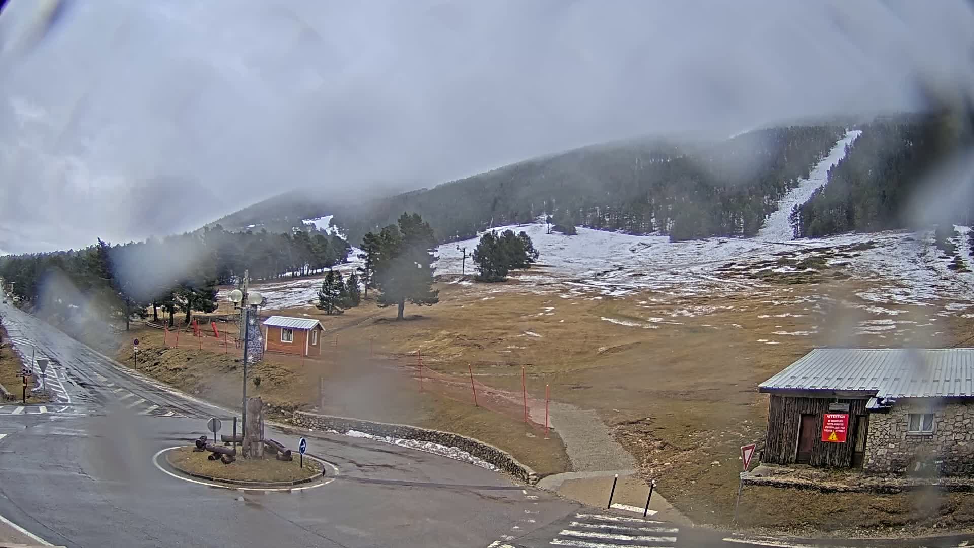 A wet, overcast view of a mountain road leading to a ski resort with some remaining snow, small buildings, and a roundabout.