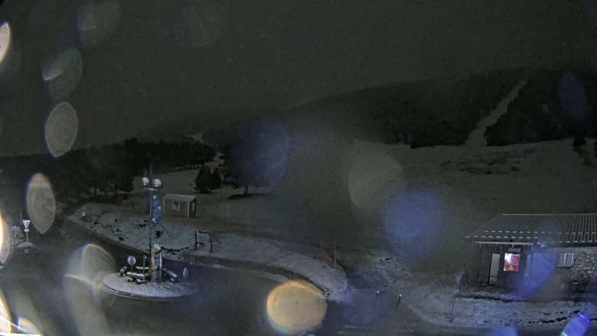 A snow-covered nighttime scene shows a small building, a roundabout, and a hillside with faint traces of a road or path.