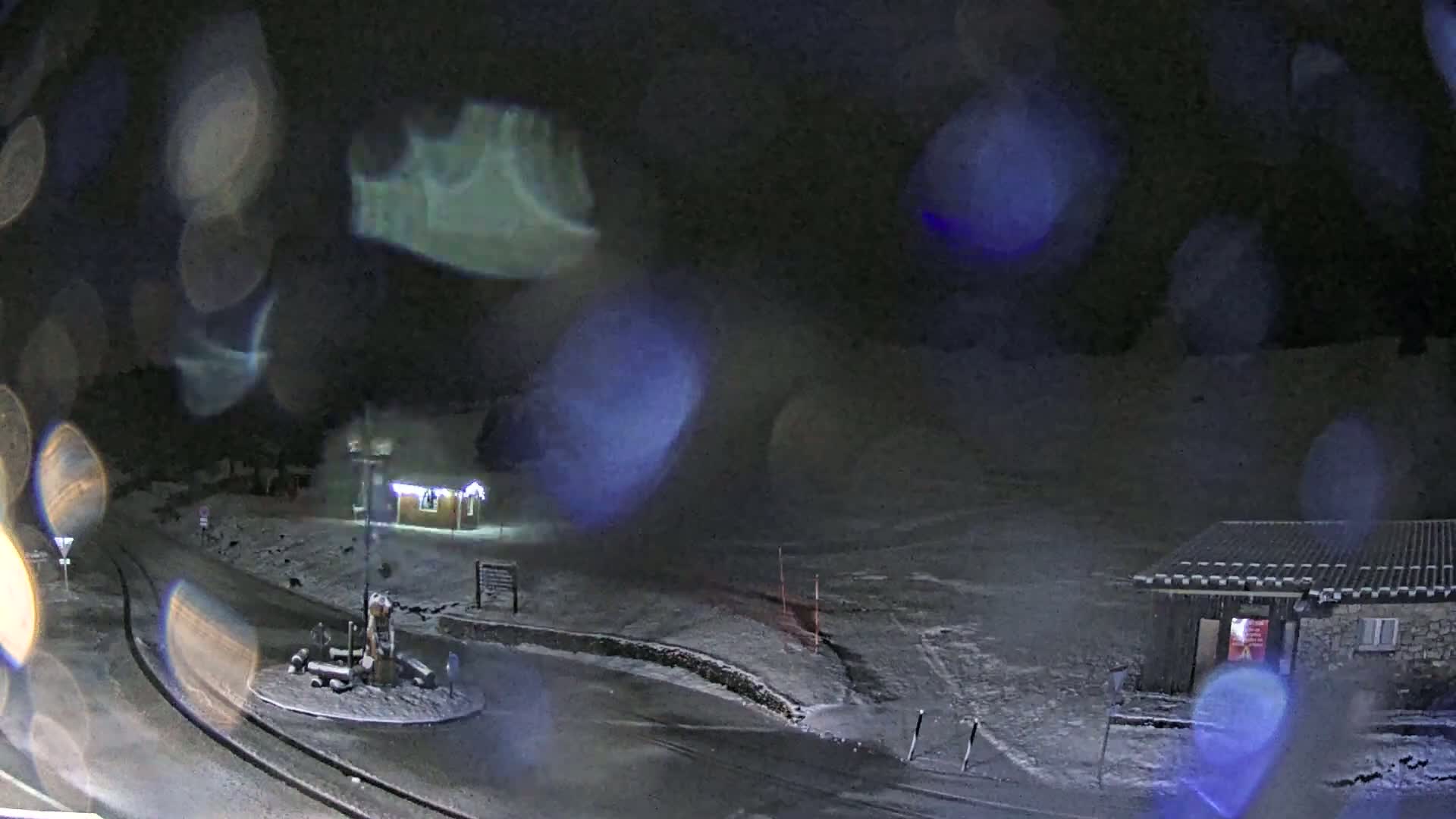 A snow-covered roadway curves around a small roundabout with a structure in the center, near small buildings under a dark, nighttime sky with blurry raindrops or snowflakes visible.