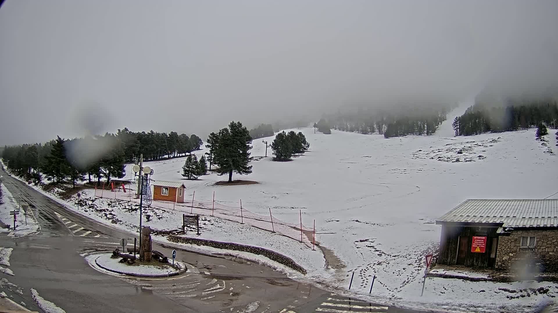 A snow-covered ski slope is visible under an overcast sky, with rain or snow falling, and a road and small buildings near the bottom of the slope.