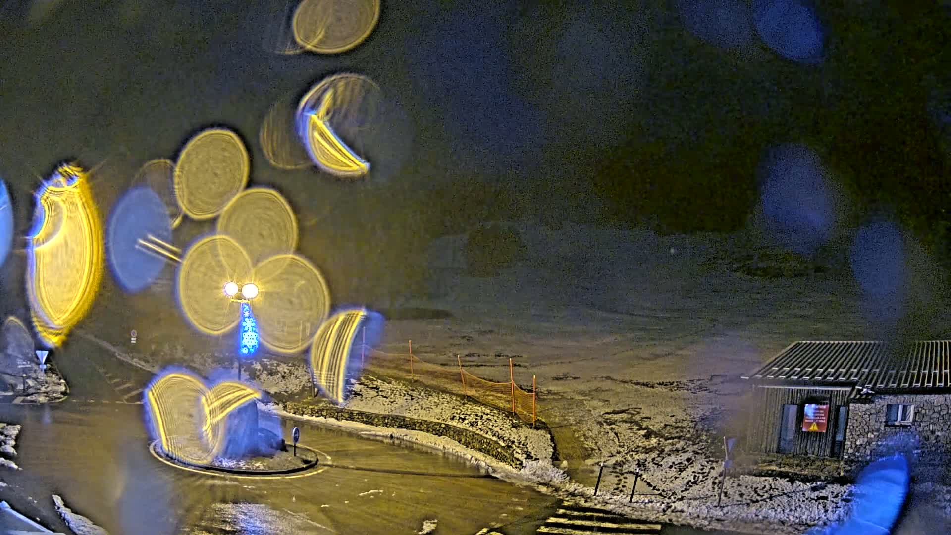 A nighttime view of a snow-covered roundabout and a small building, seen through rain-streaked glass producing blurred light streaks.