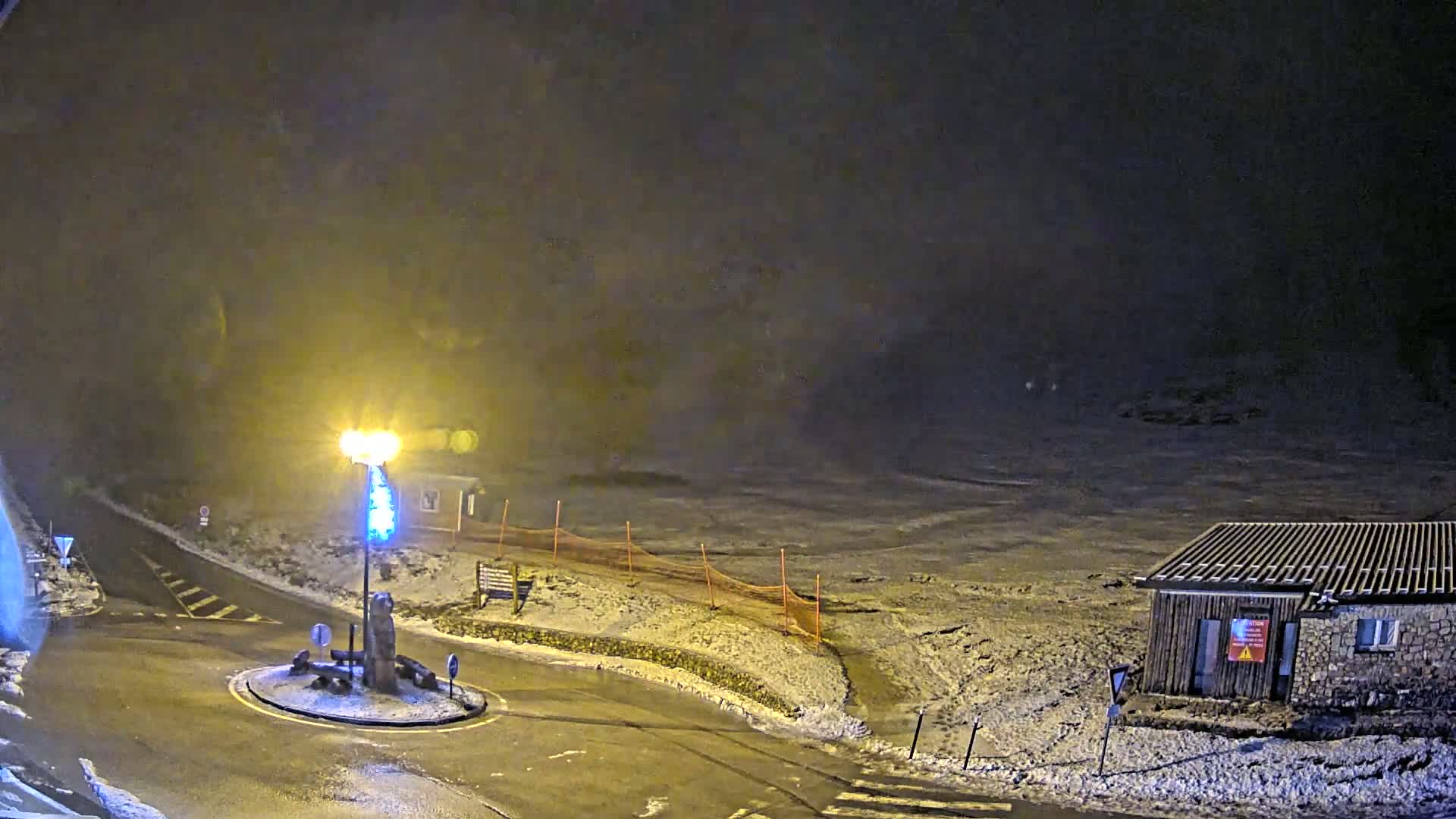 A nighttime scene of a snow-covered road intersecting a roundabout with a light pole and small building, under hazy conditions.