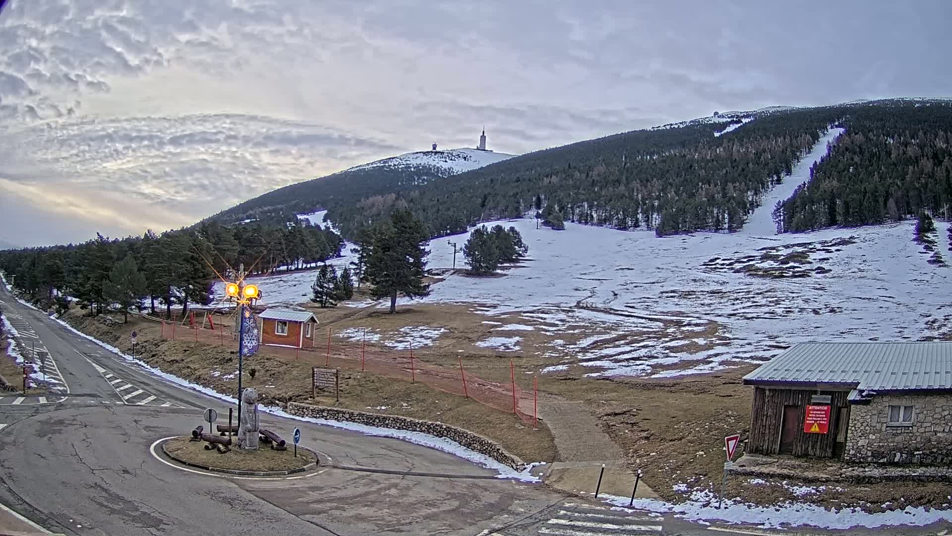 A partially snow-covered mountain slope with a tall tower on its peak is visible under a cloudy sky, with a road and small buildings in the foreground.