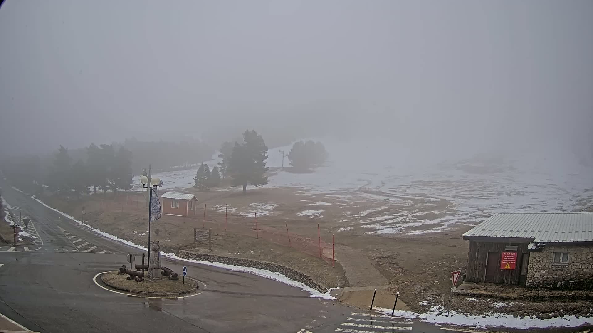 A foggy, snow-dusted landscape features a road intersection, several small buildings, and scattered trees.