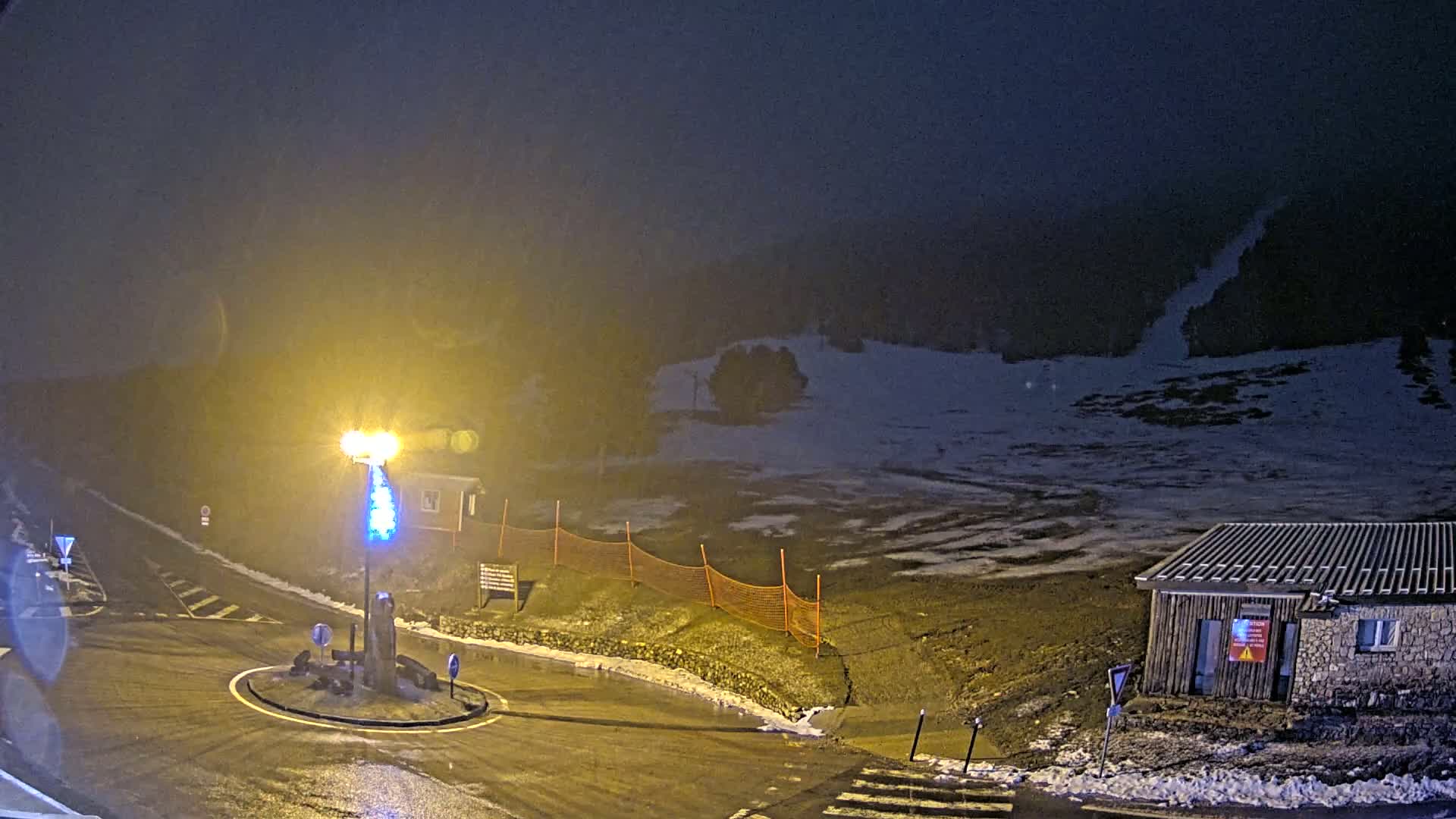 A nighttime, snow-covered scene shows a roundabout and a small building near a ski slope under hazy conditions.