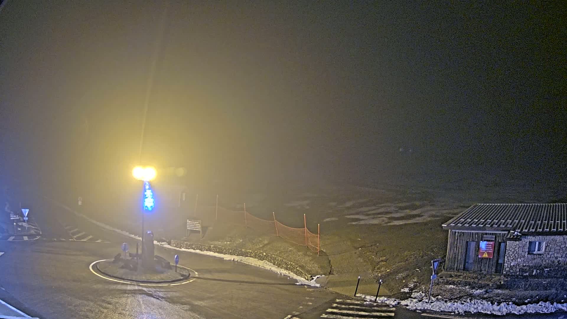 A foggy night scene shows a small building and a roundabout at the intersection of roads, partially covered in snow.