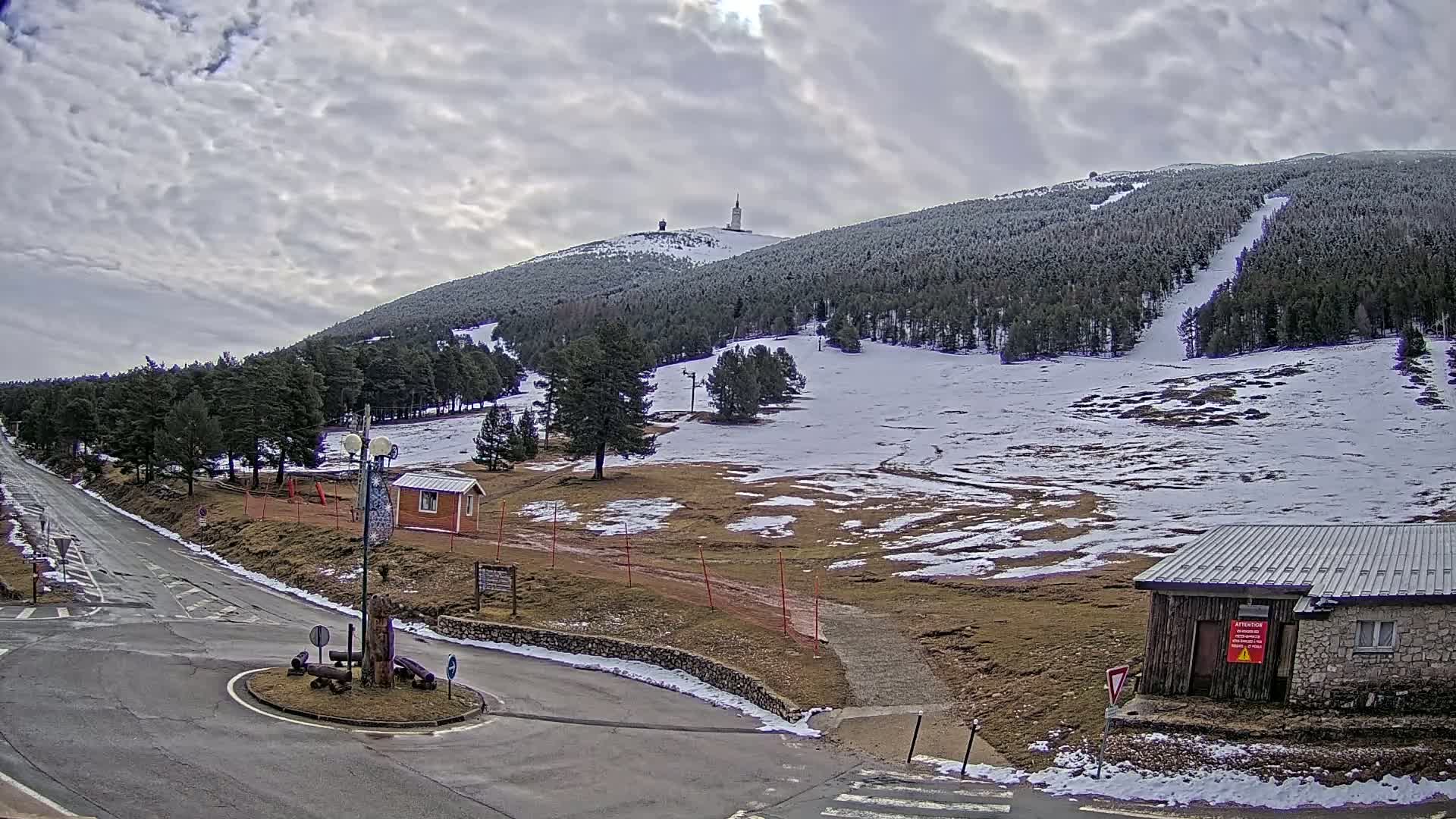 A snow-dusted mountain slope with a tall tower at its peak is viewed from a road intersection, under a cloudy sky.