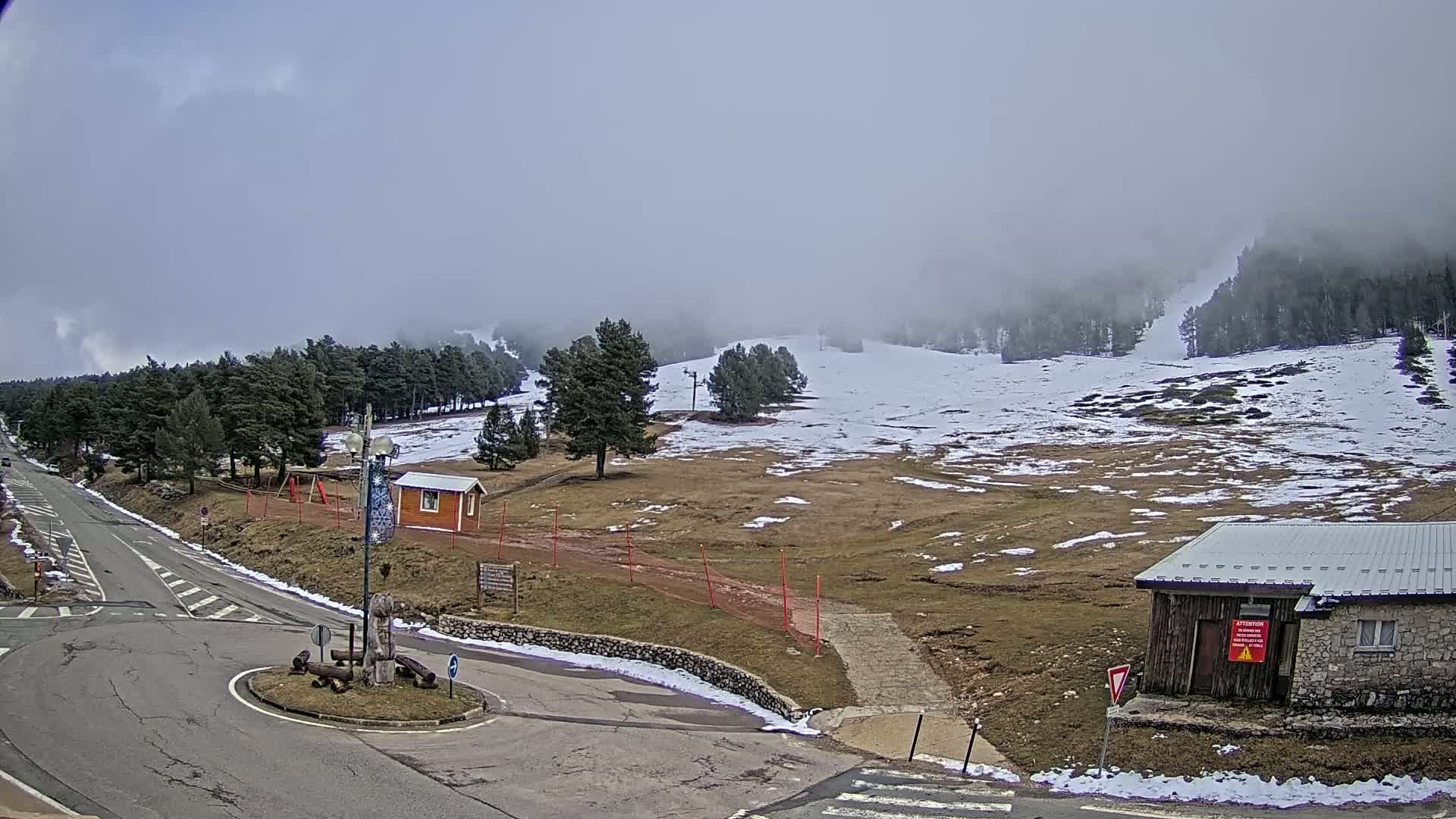 A mostly cloudy day shows a snowy, hilly landscape with a road leading to a small building near a roundabout.