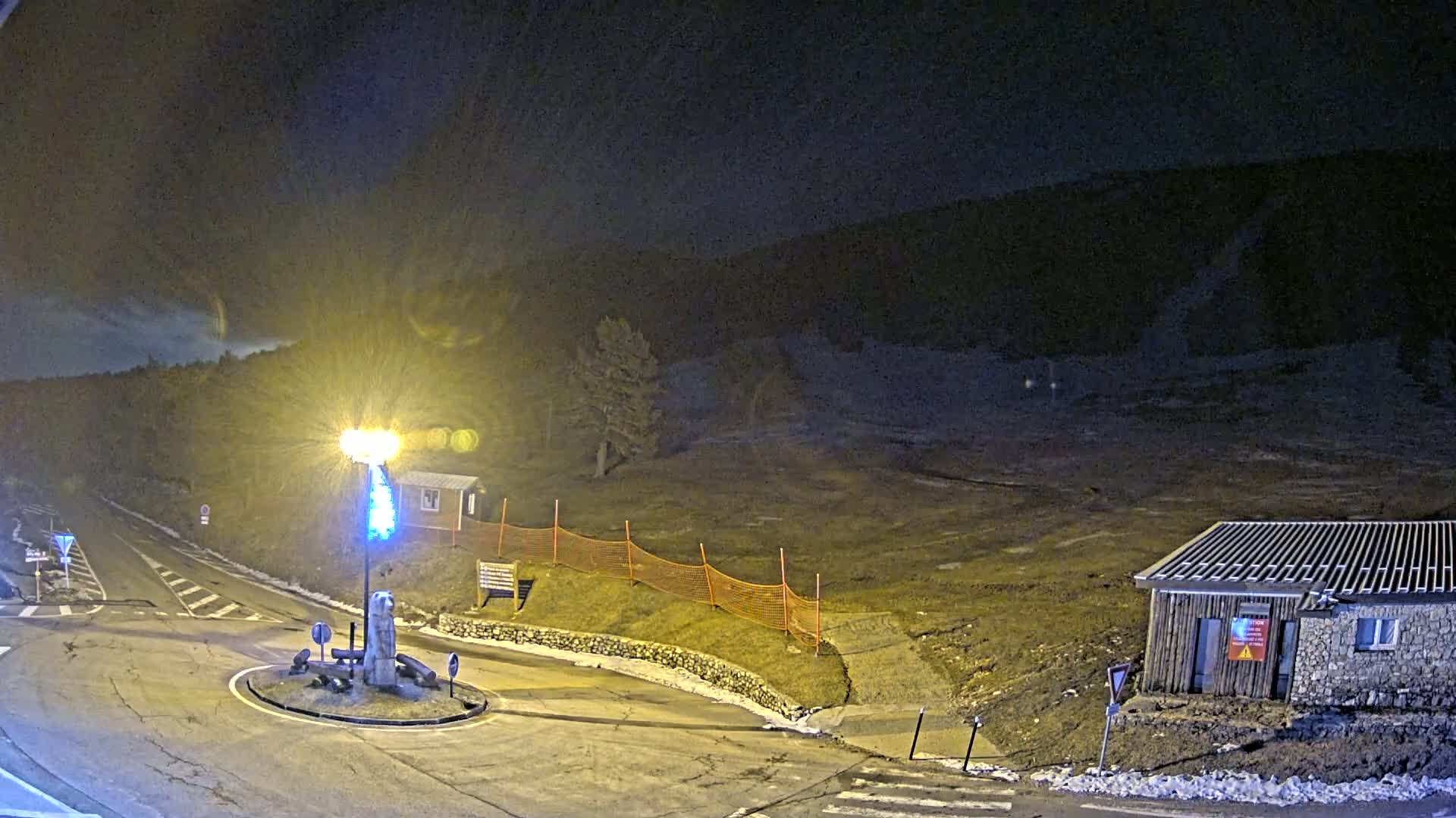 A nighttime view of a roundabout and a small building near a hill, with some snow on the ground and an orange safety fence.