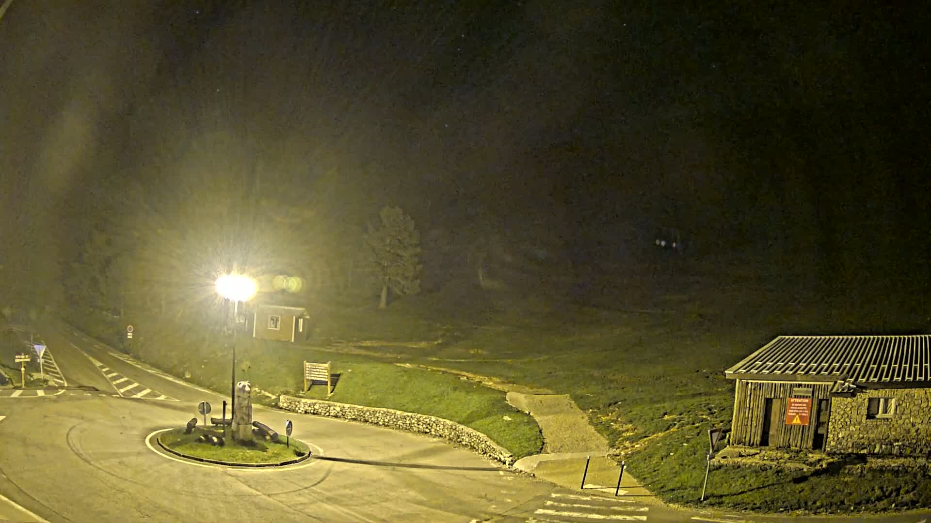 A nighttime view of a roundabout and small buildings illuminated by a single lamppost, with a dark, grassy area extending into the background under a mostly clear night sky.