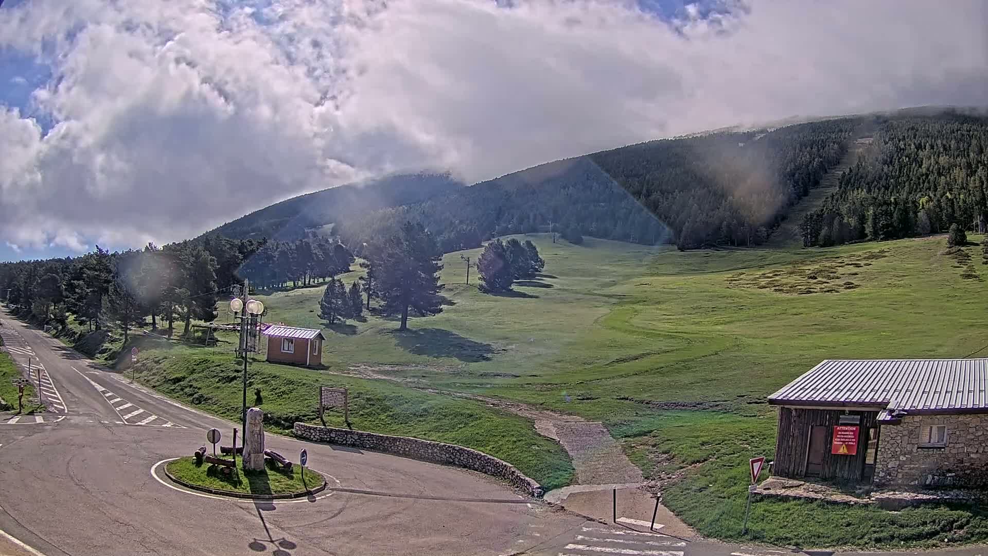 A partly cloudy day reveals a mountain road curving through a grassy plateau, flanked by small buildings and coniferous trees, with a mountain range in the background.