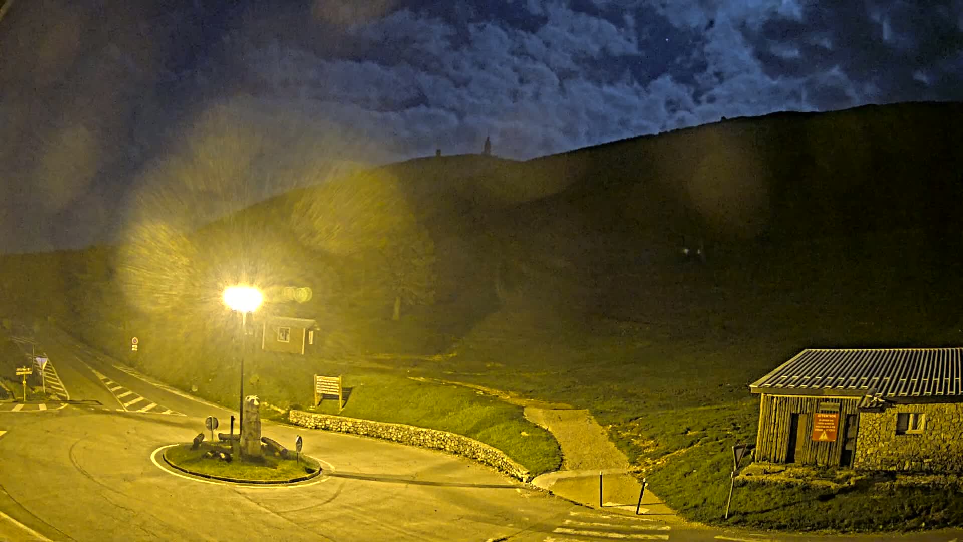 A nighttime view of a roundabout and small building near a hill under a cloudy sky.