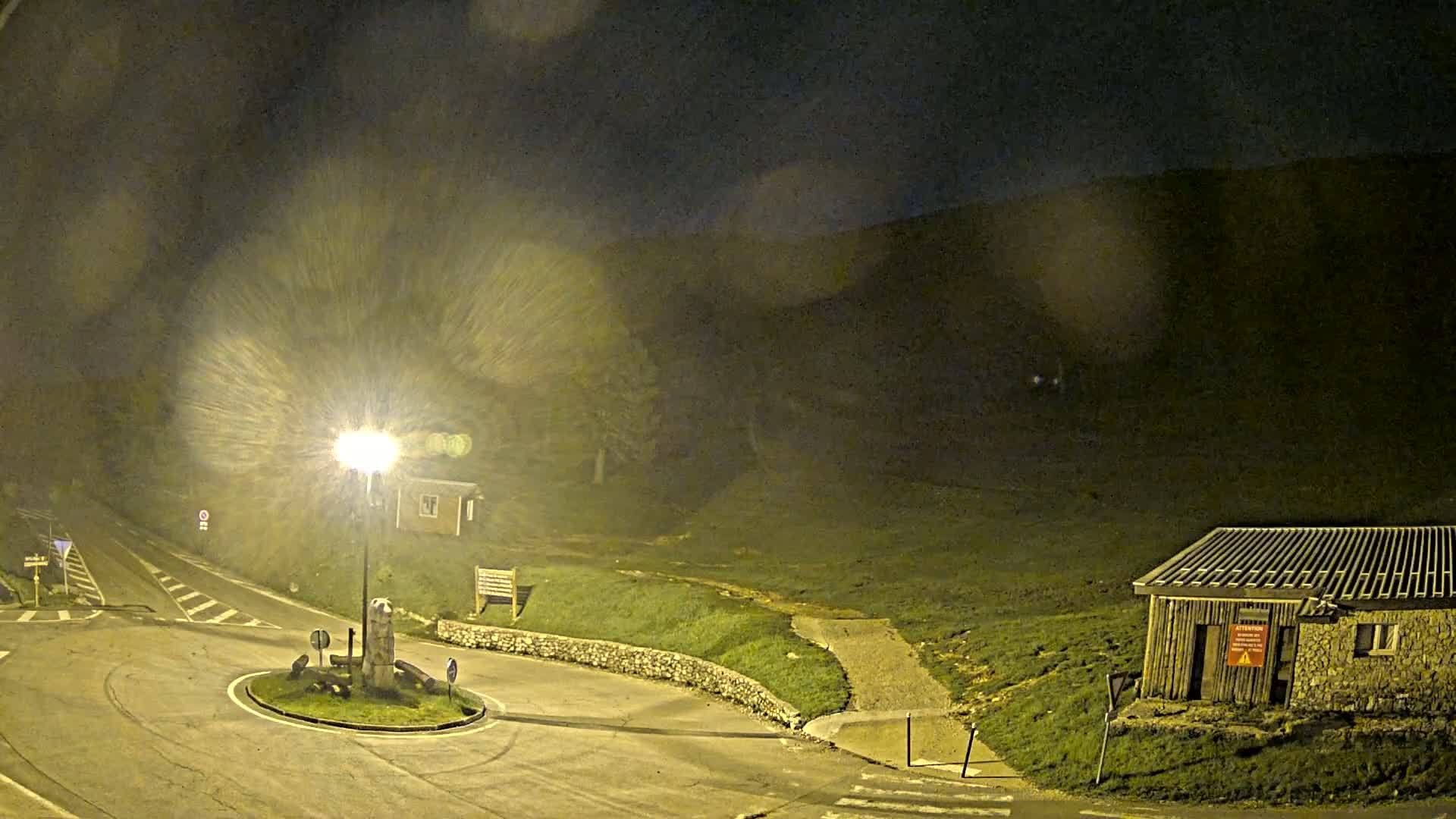 At night, under a dark, possibly rainy sky, a brightly lit roundabout sits near a small stone building and a grassy hillside.