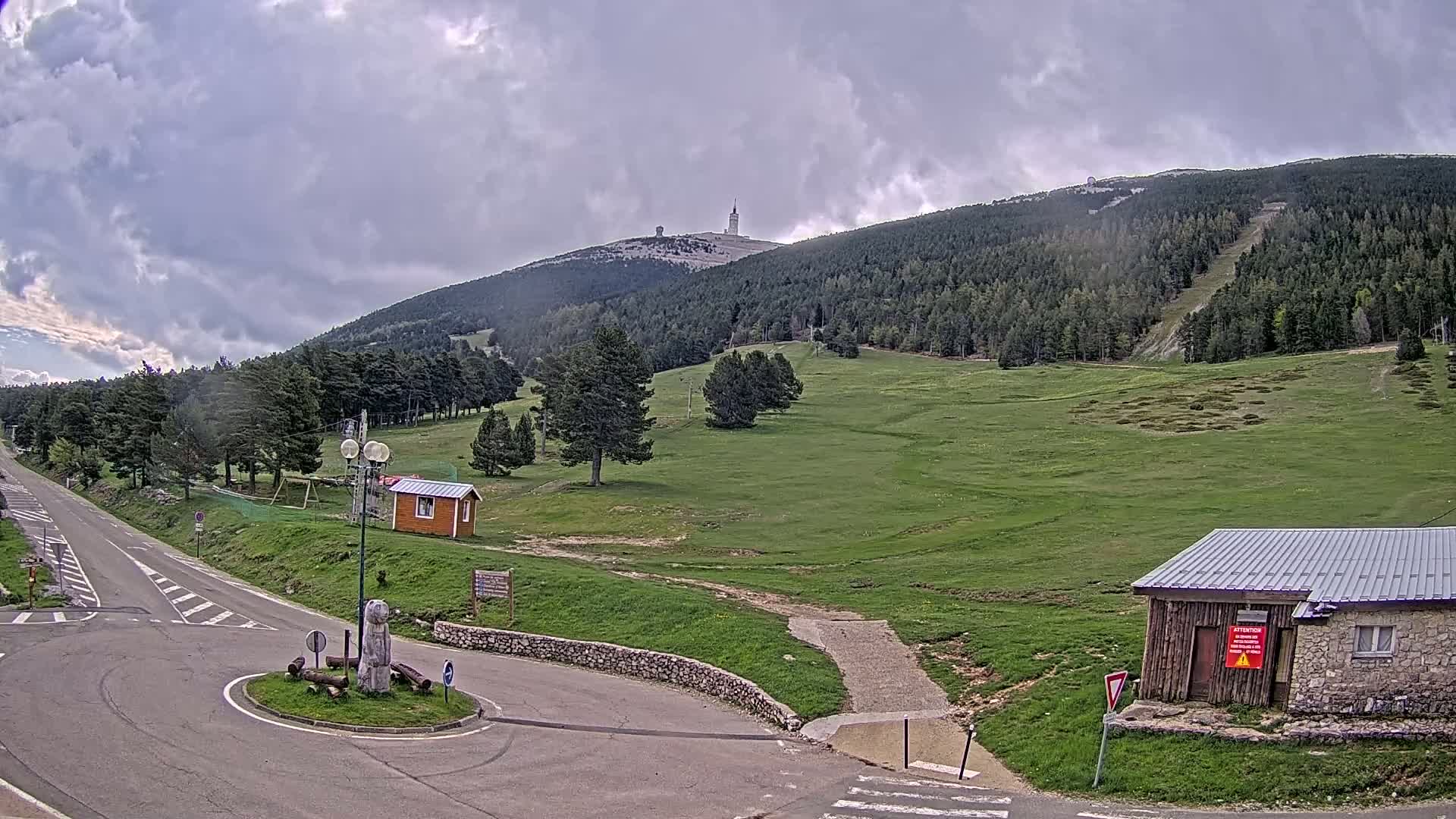 A mountaintop view shows a paved road leading to a grassy area with a few small buildings, under a cloudy sky.