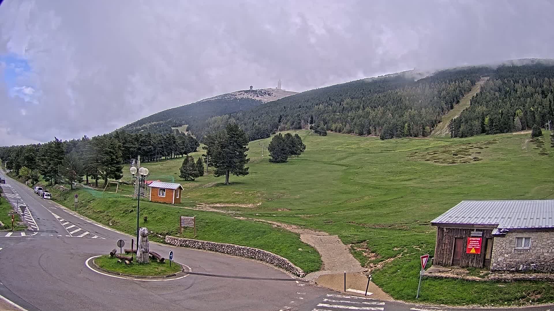 A paved road leads to a roundabout in a mountainous area with overcast skies, where green fields and forests stretch towards a distant peak with a structure on top.