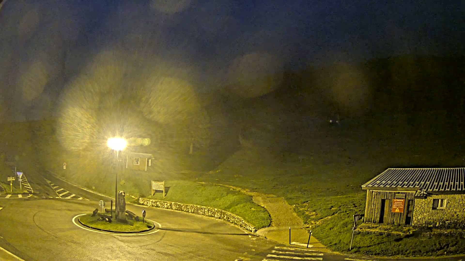 A nighttime scene of a roundabout with a small building and a bright streetlight, illuminated under heavy rain or snow.