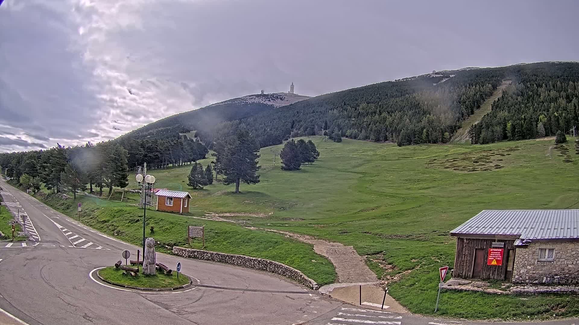 A paved road leads to a grassy mountain plateau under a cloudy sky, with small buildings and a distant tower visible on the mountainside.