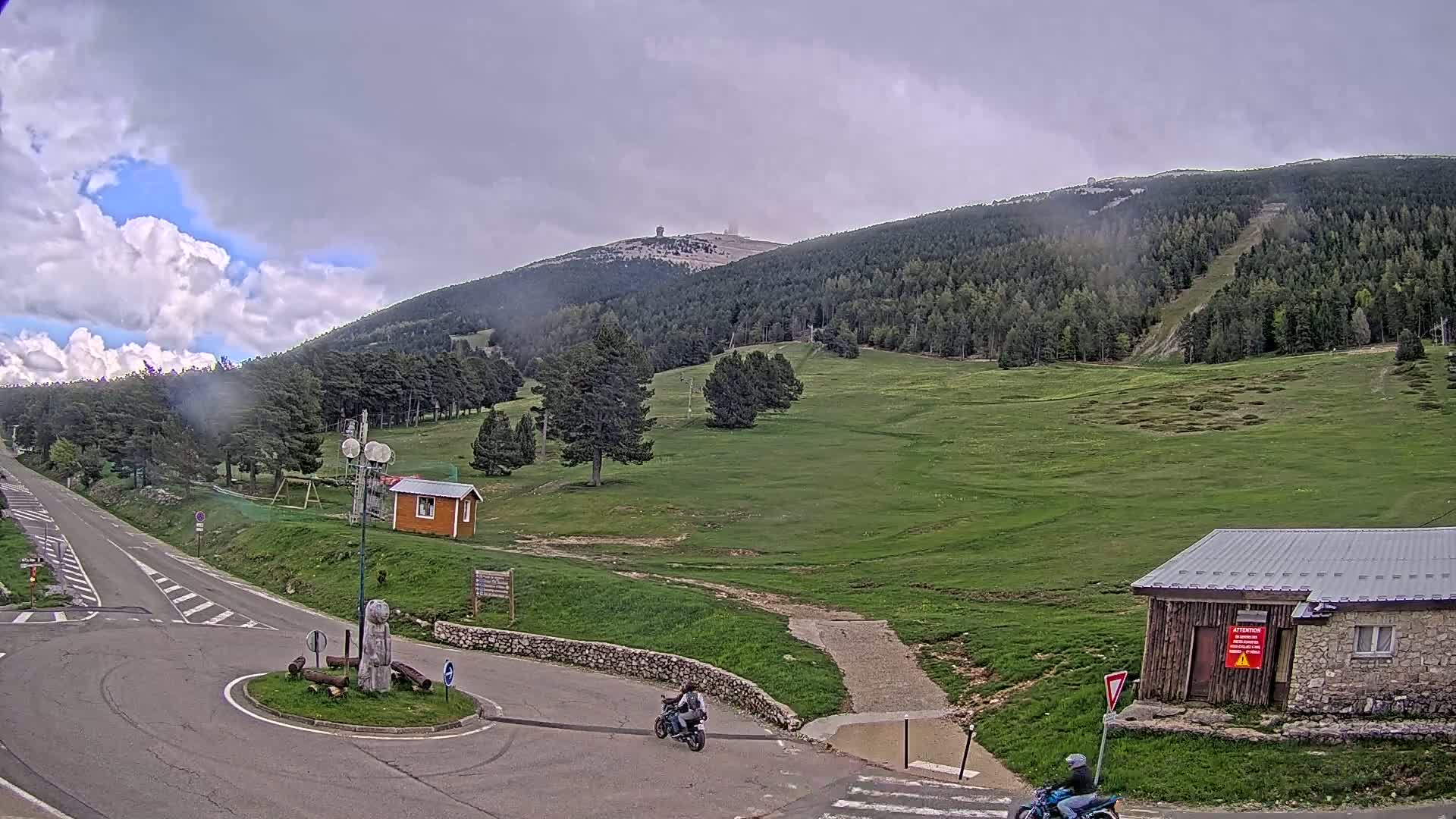 A partly cloudy sky hangs over a mountainous landscape with green meadows, scattered trees, small buildings, and a road with motorcycles.