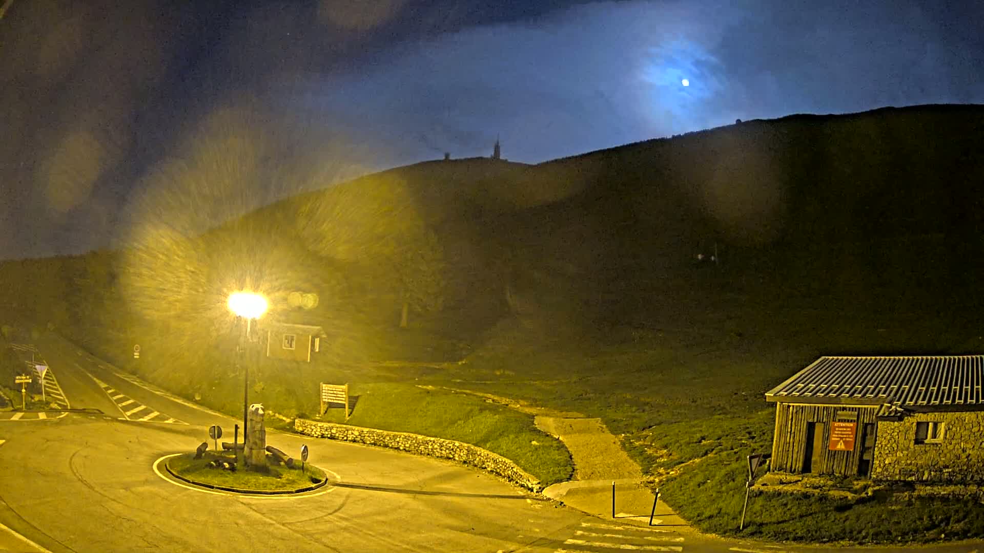 A moonlit night scene shows a roundabout and a small building at the base of a hill under rainy conditions, with a light post illuminating the area.