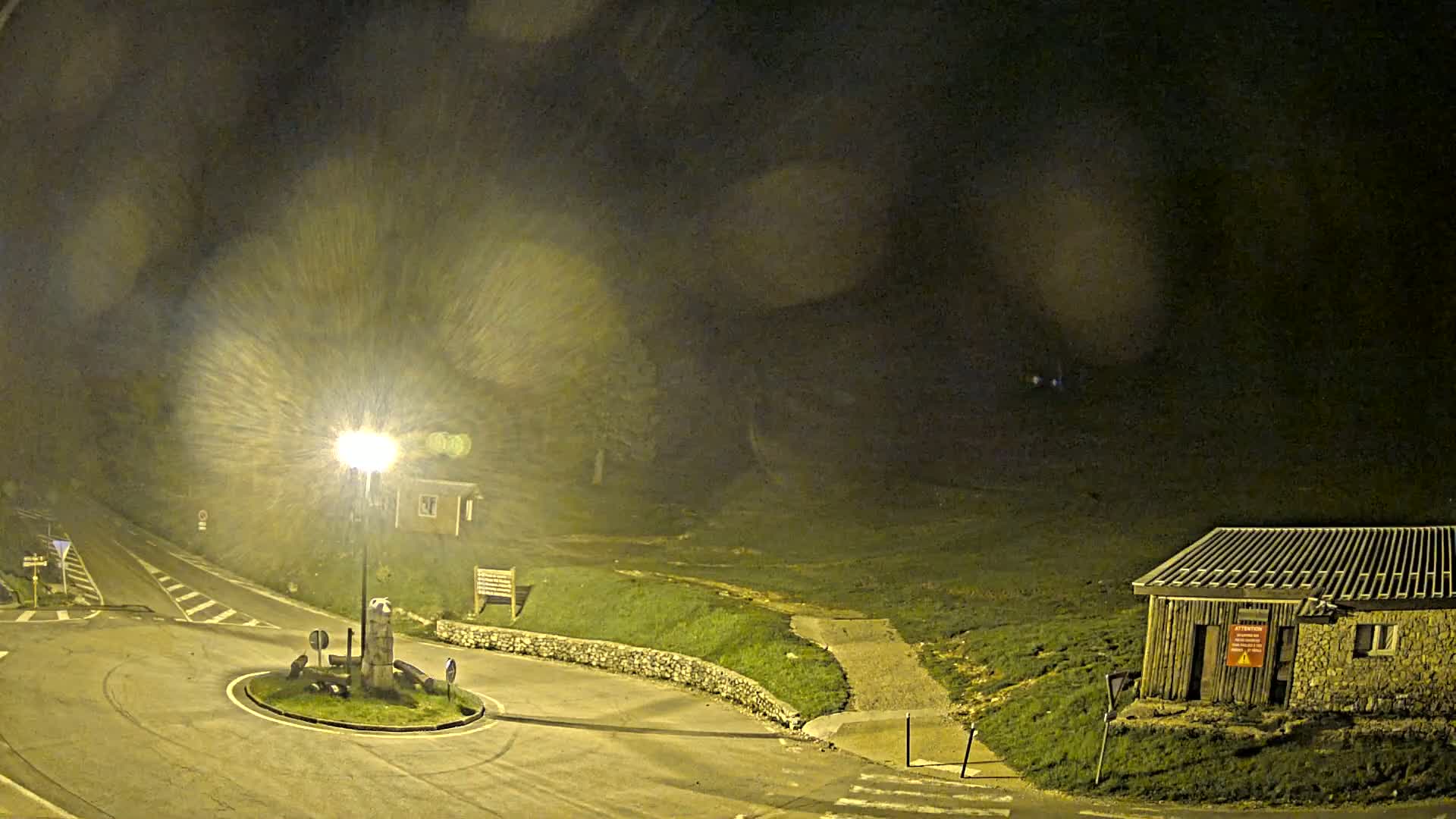 A nighttime scene shows a roundabout with a small building and a street light in light snowfall.
