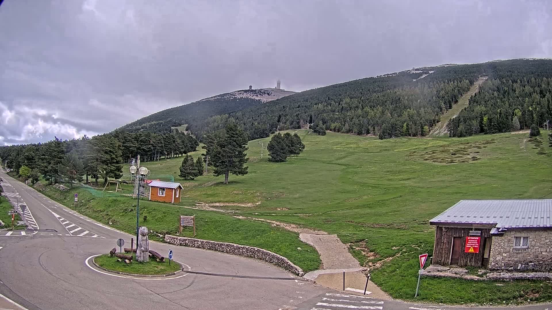 A paved road leads to a grassy area with small buildings, a stone sculpture, and a mountain in the background under a cloudy sky.