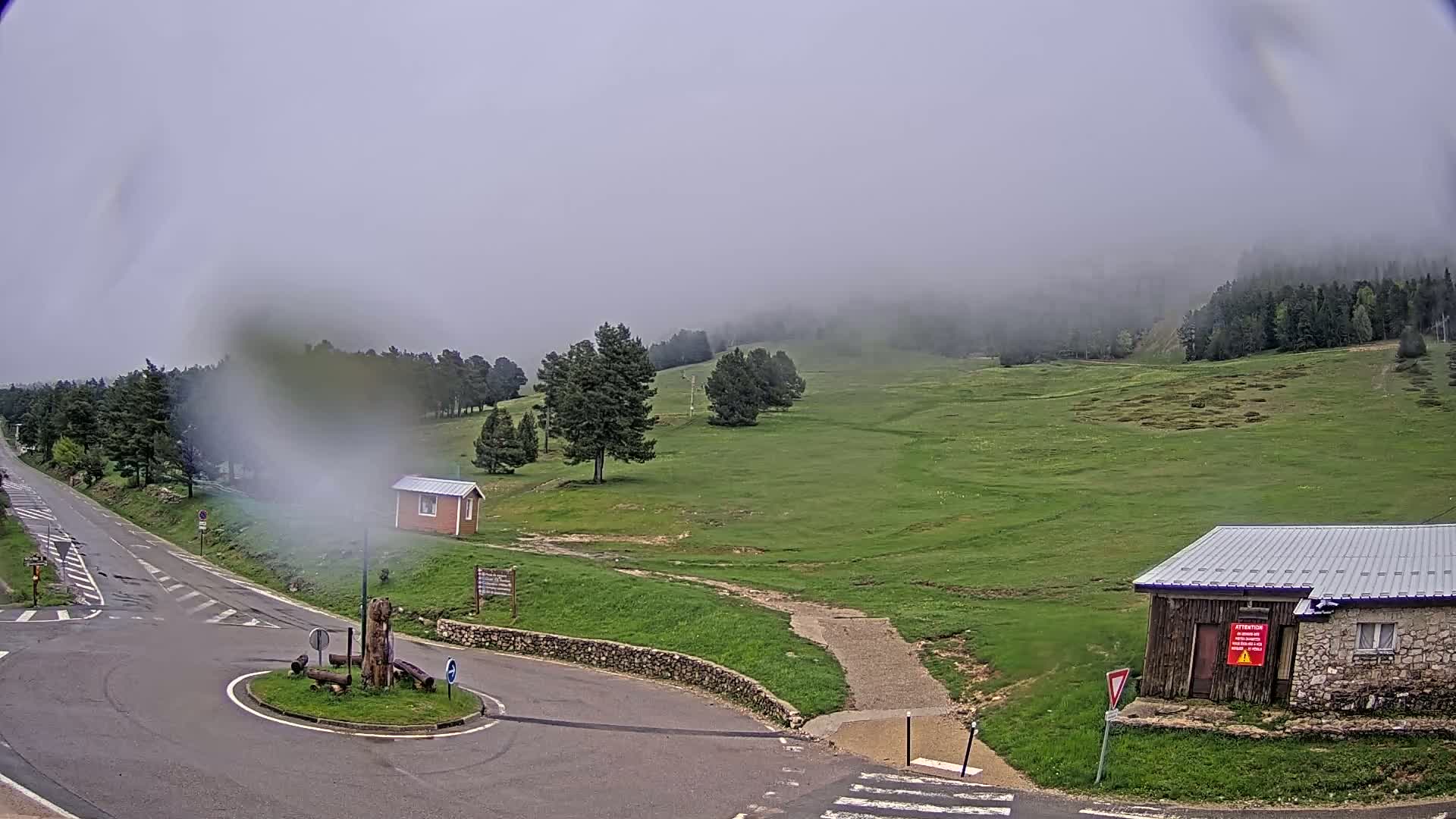 A foggy, overcast day reveals a rural scene with a road intersection, small buildings, and a grassy hillside dotted with trees.