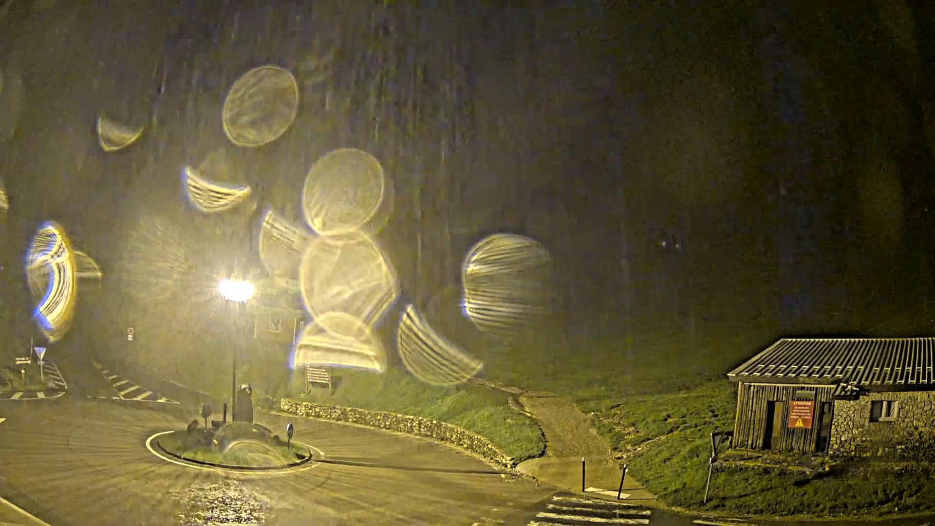 A nighttime scene shows a wet roundabout with a streetlight, a small building, and numerous blurry, circular light streaks in the sky suggesting heavy rain or perhaps lens distortion.