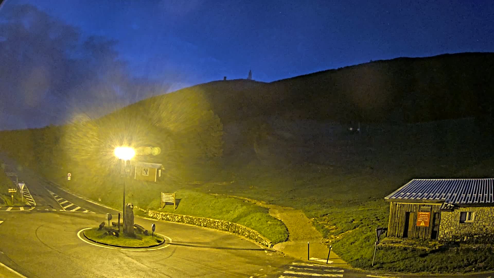 A nighttime scene shows a roundabout and a small building next to a hill under a dark blue, cloudy sky illuminated by a bright streetlight.