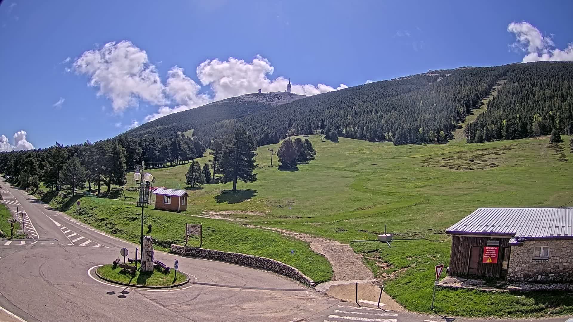 A mountaintop with a tall tower is visible in the distance under a partly cloudy, sunny sky, with a grassy field and roadway in the foreground.