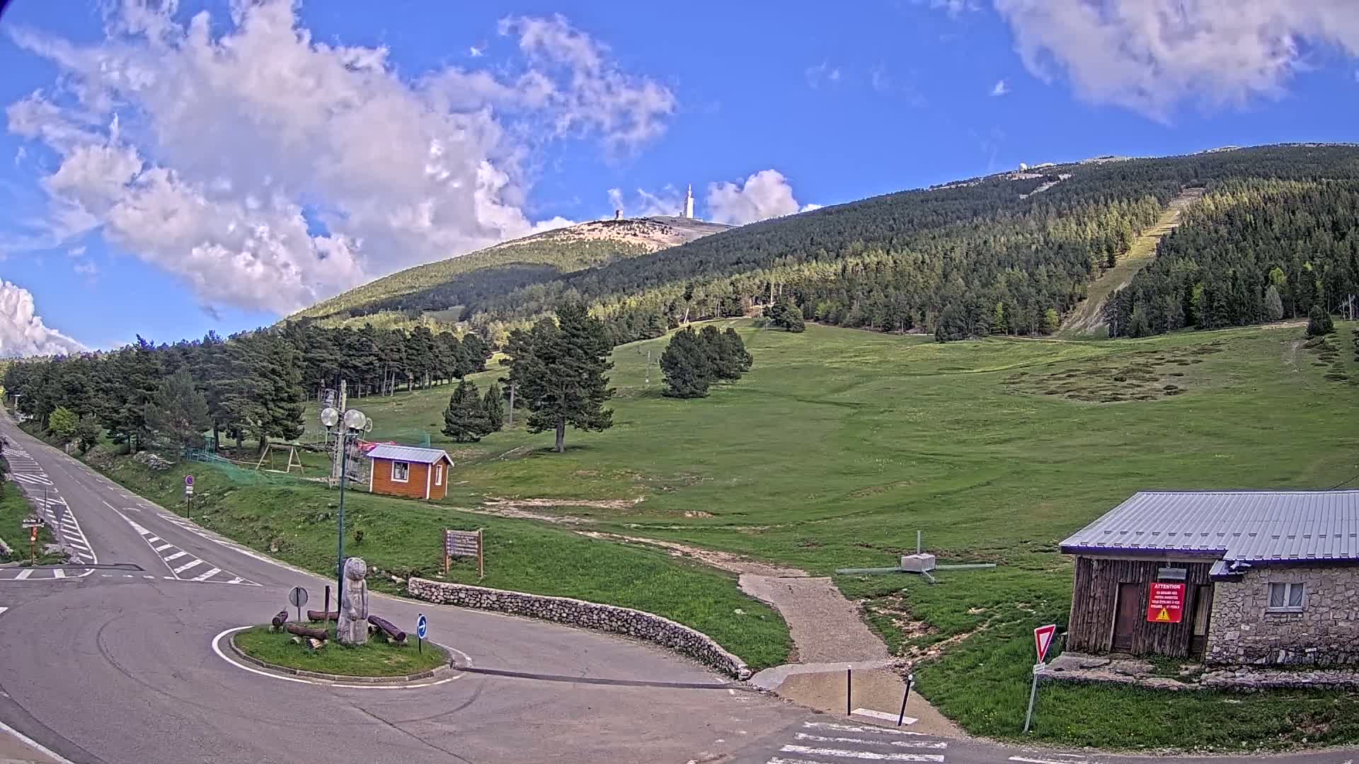 A mountaintop with a communications tower is visible in the distance under a partly cloudy sky, with a grassy area and small buildings at the base of the mountain near a roadway.