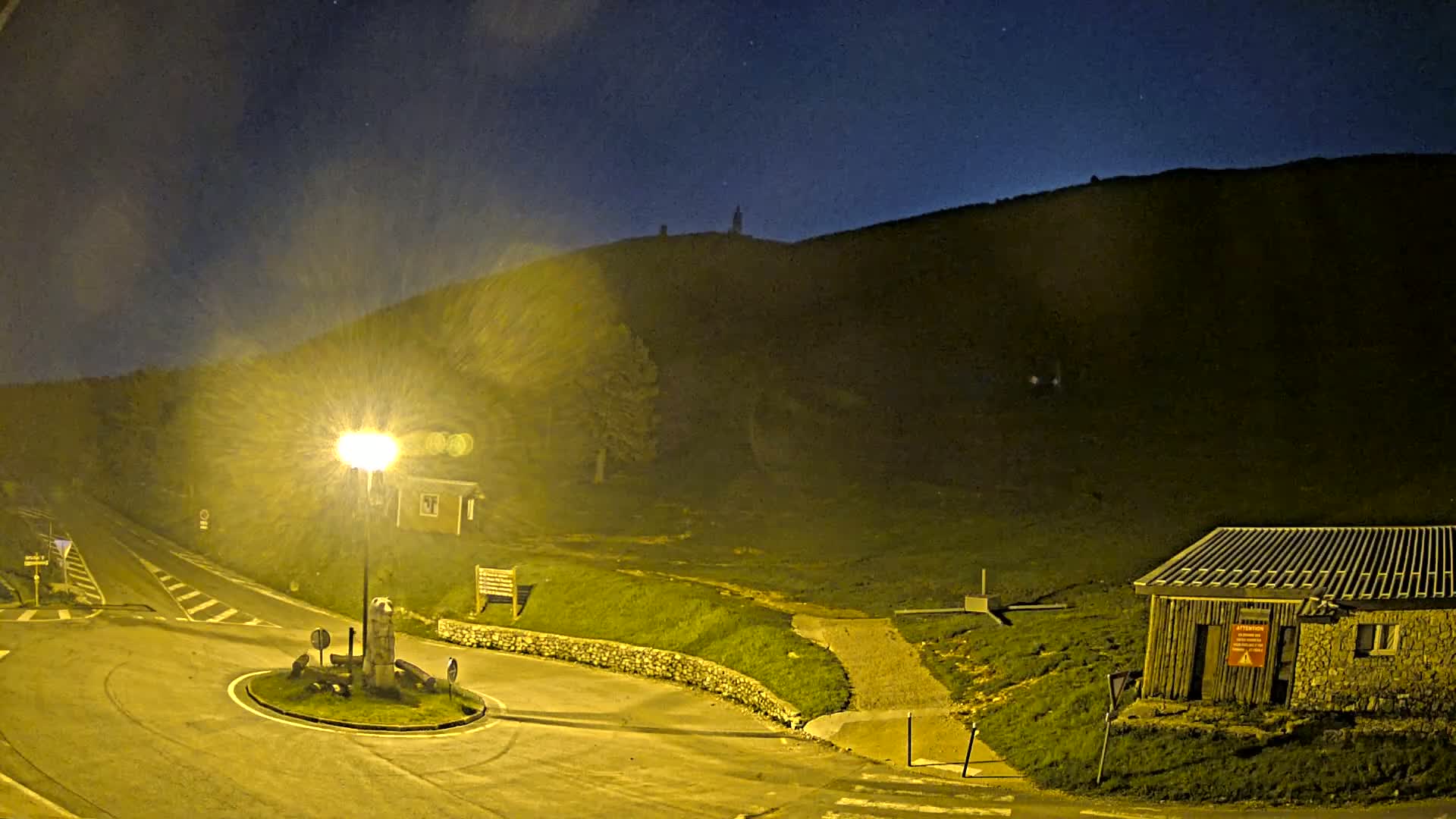 A nighttime view of a roundabout and small building illuminated by a lamppost, with a dark hill rising in the background under a slightly hazy night sky.