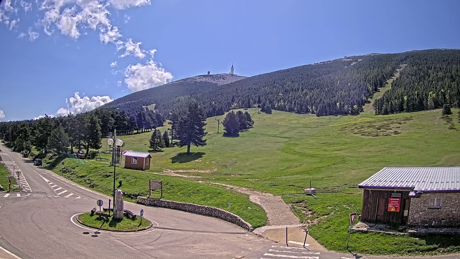 A paved road leads to a grassy mountainside with a tall tower atop a forested peak under a mostly sunny sky with some clouds.