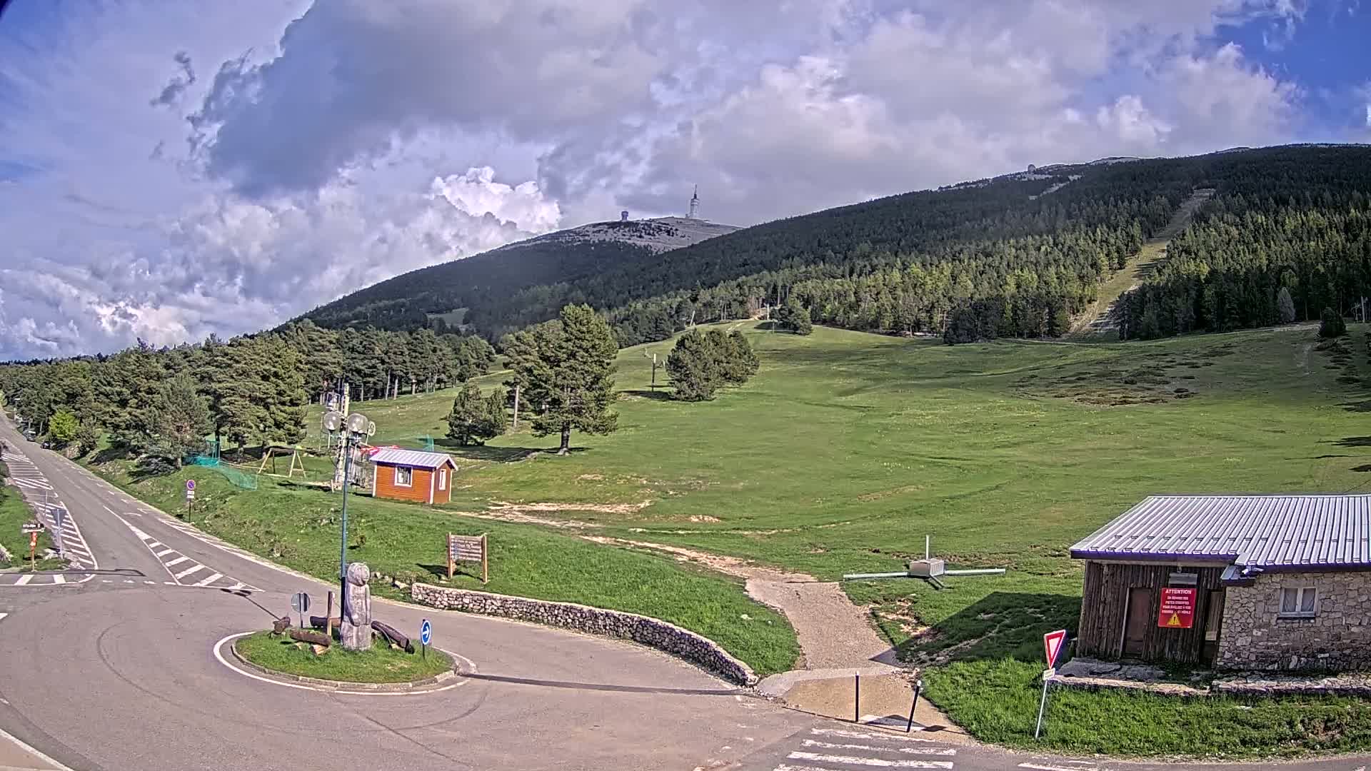 A paved road curves around a grassy area with small buildings near a mountain covered in green trees under a partly cloudy sky.