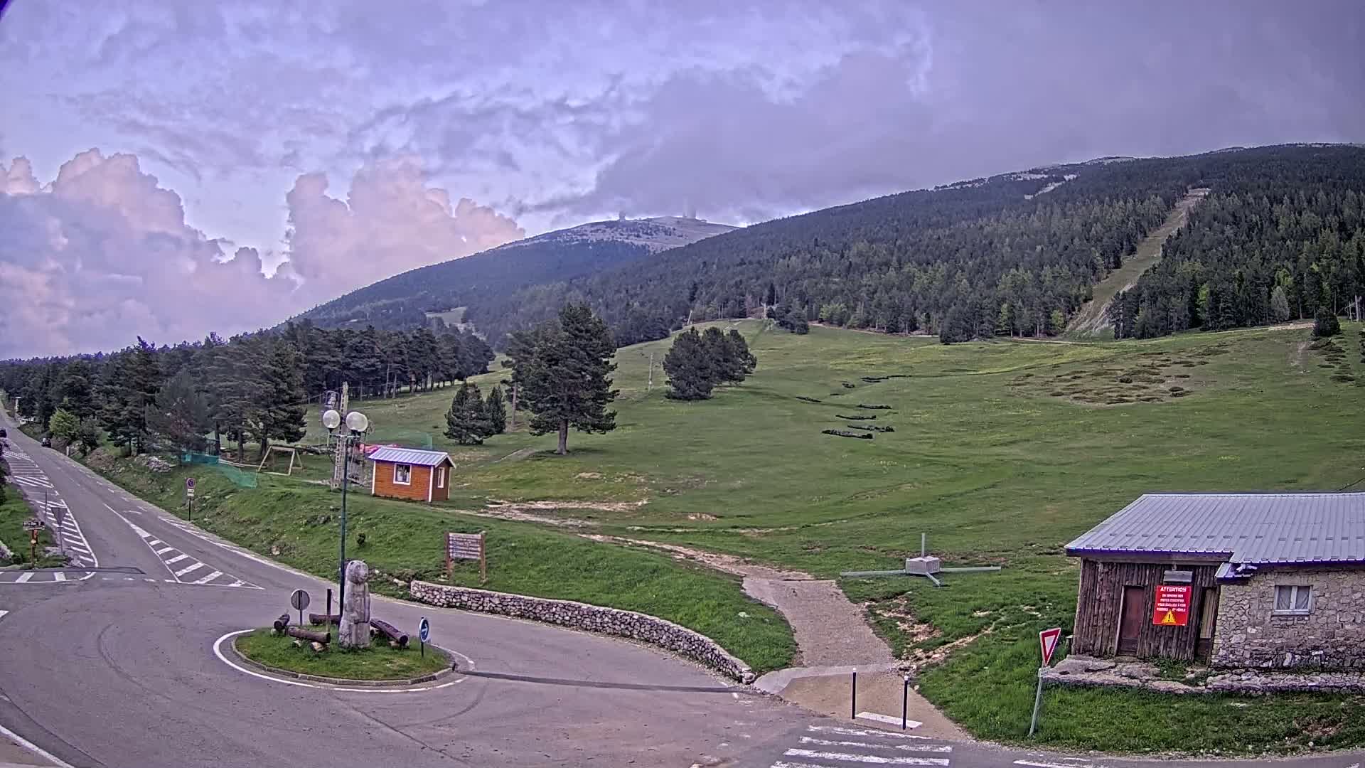 A mountain road intersects a grassy area with small buildings, under a partly cloudy sky.