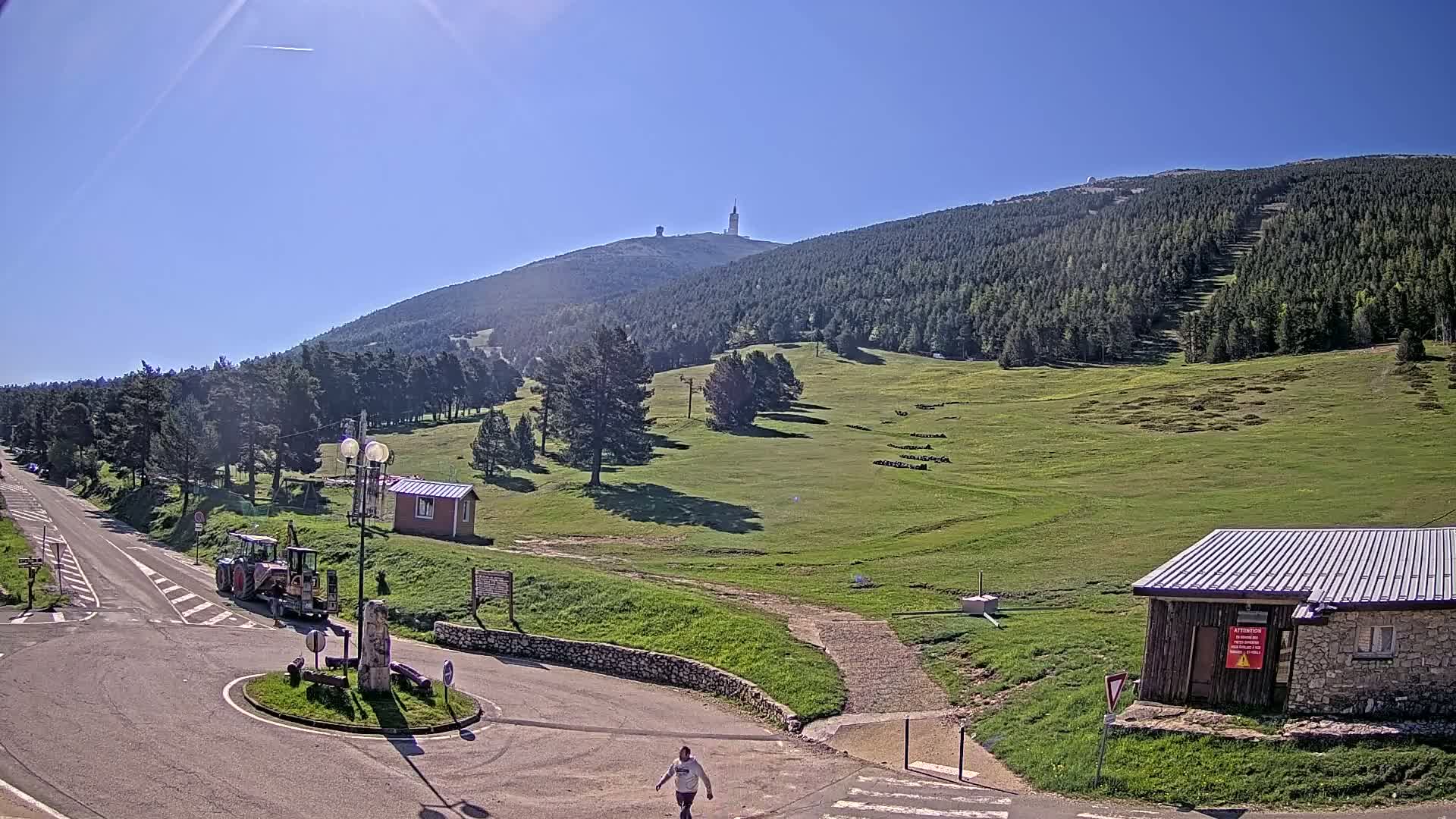 A sunny mountain vista reveals a grassy hillside with sparse trees, a road leading to a small building, and a tall tower atop a distant mountain peak.